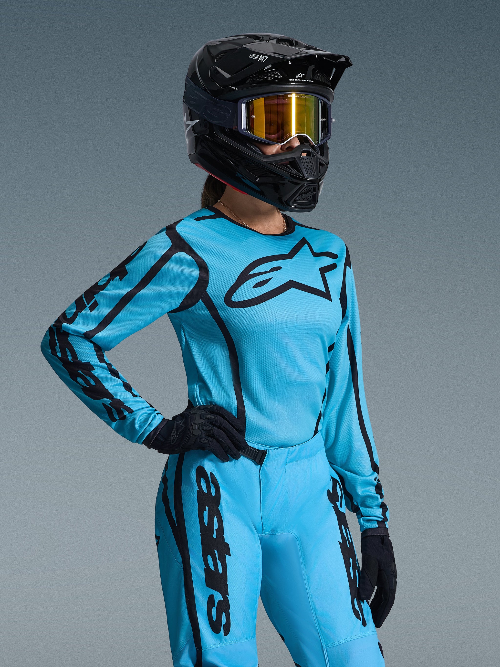 Motocross rider wearing the Alpinestars 2026 Woman Stella Fluid Apex Jersey in Tropical Black, bright cyan jersey with black logos, matching pants, black helmet with orange-tinted goggles, and black gloves, professional off-road gear set for women.