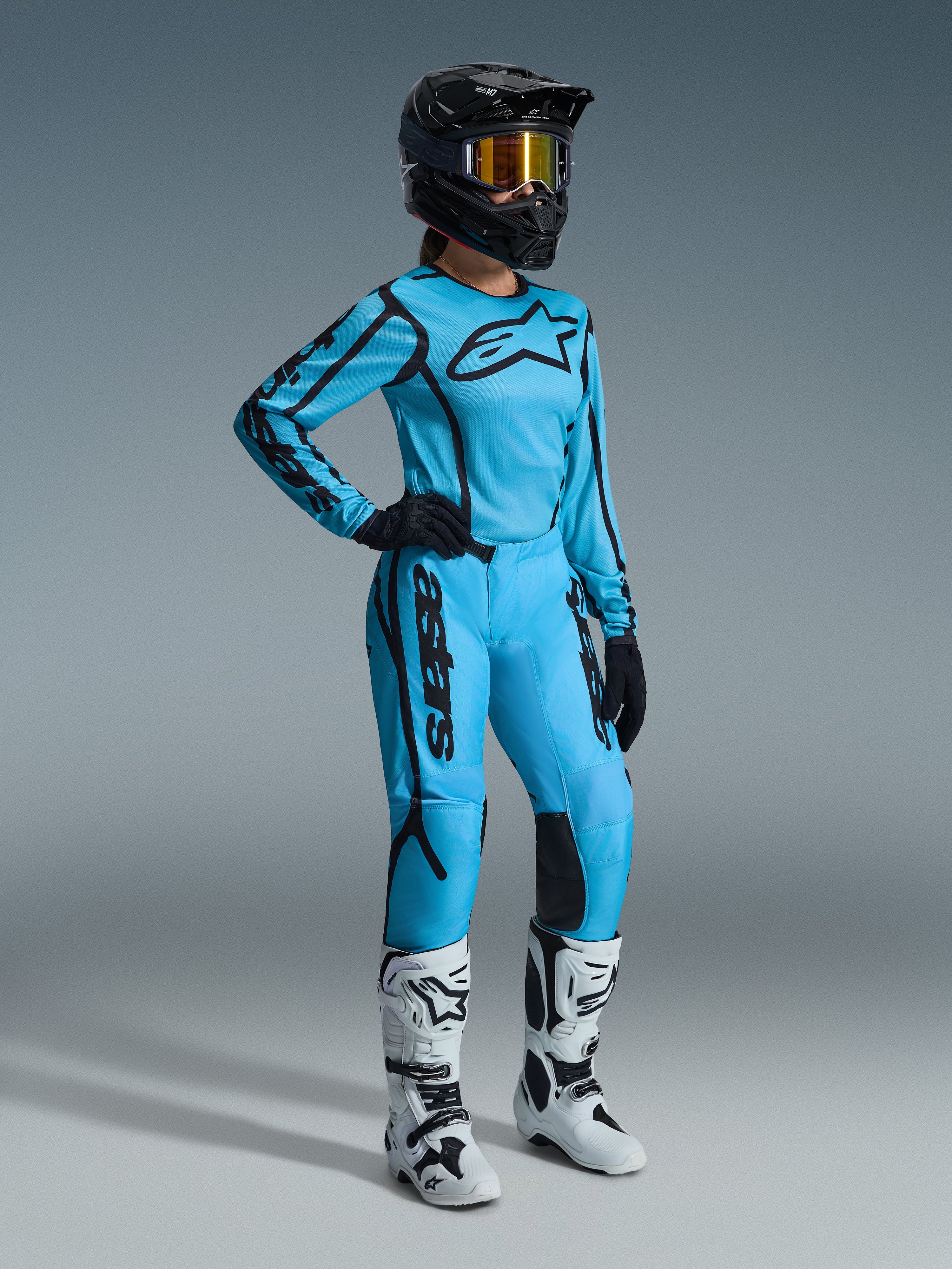 Alpinestars 2026 Stella Fluid Apex utrustningsset för dam i Tropical Black, bestående av en ljusblå tröja och matchande byxor med svarta detaljer, bärs av en kvinnlig förare med svart hjälm, iriserande goggles och vita stövlar, professionell motocrossutrustning.