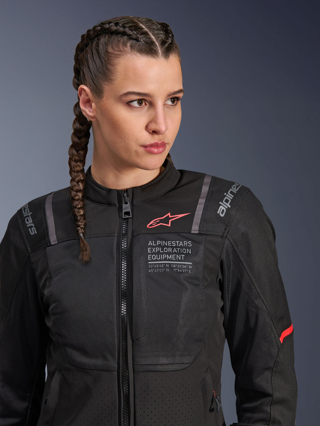 Alpinestars Stella ST-2 Air Jacka, MC-jacka för dam i svart, med meshpaneler för luftflöde och 'Alpinestars Exploration Equipment'-märkning med koordinater, röd logotypdetalj på bröstet, bärs av en modell med flätat hår.