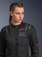 Alpinestars Stella ST-2 Air Jacka, MC-jacka för dam i svart, med meshpaneler för luftflöde och 'Alpinestars Exploration Equipment'-märkning med koordinater, röd logotypdetalj på bröstet, bärs av en modell med flätat hår.