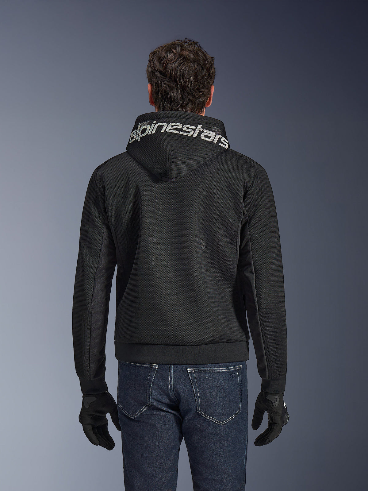 Alpinestars Chrome Superairflow Hoodie, svart motorcykelhoodie, baksida med tydlig vit Alpinestars-logotyp över huvan, teknisk 3D-meshkonstruktion, bärs med mörkblå jeans och svarta handskar, ventilerad urban mc-utrustning