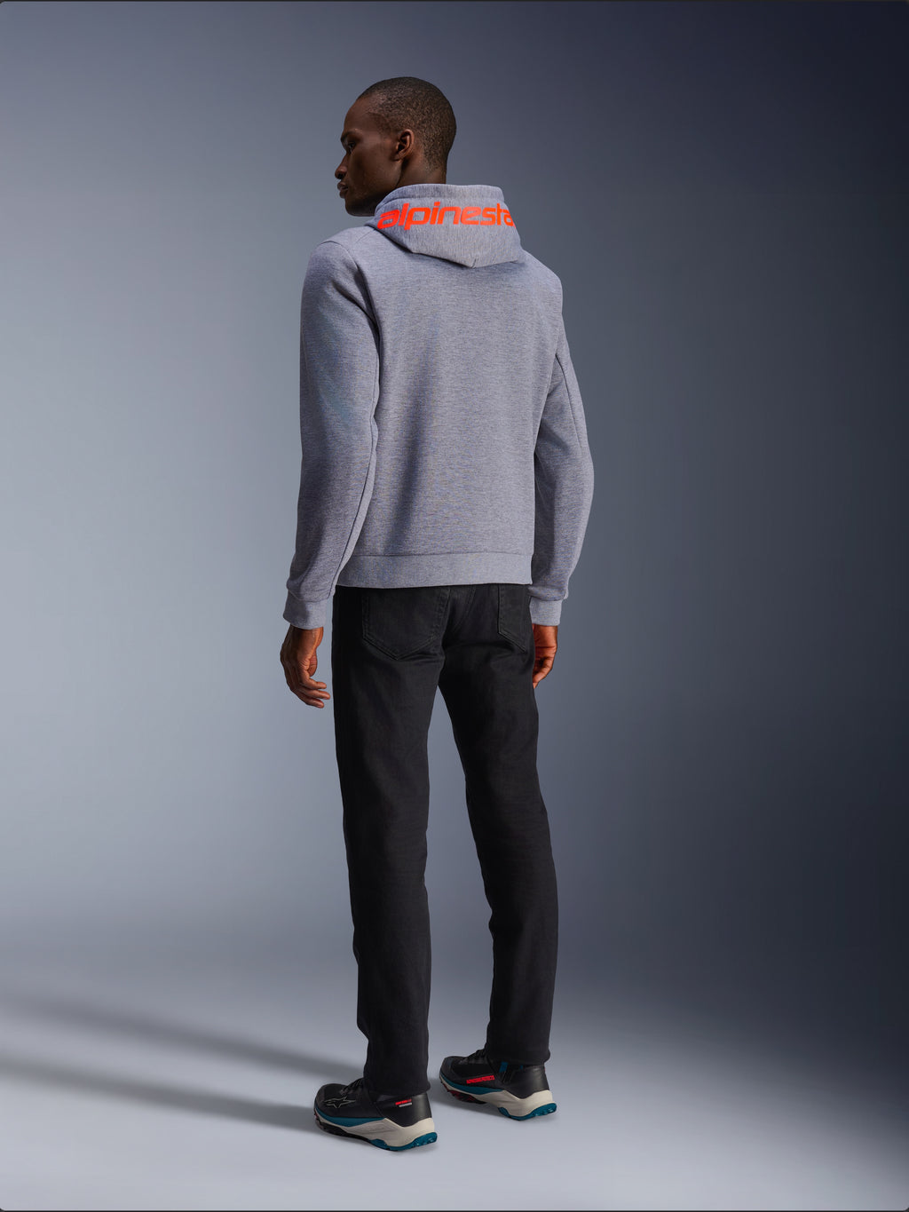 Alpinestars Chrome V2 Sport Hoodie in Gray Heather, back view showing a large Red Fluo Alpinestars wordmark across the hood, worn by a model with black pants and dark riding shoes, casual urban motorcycle apparel