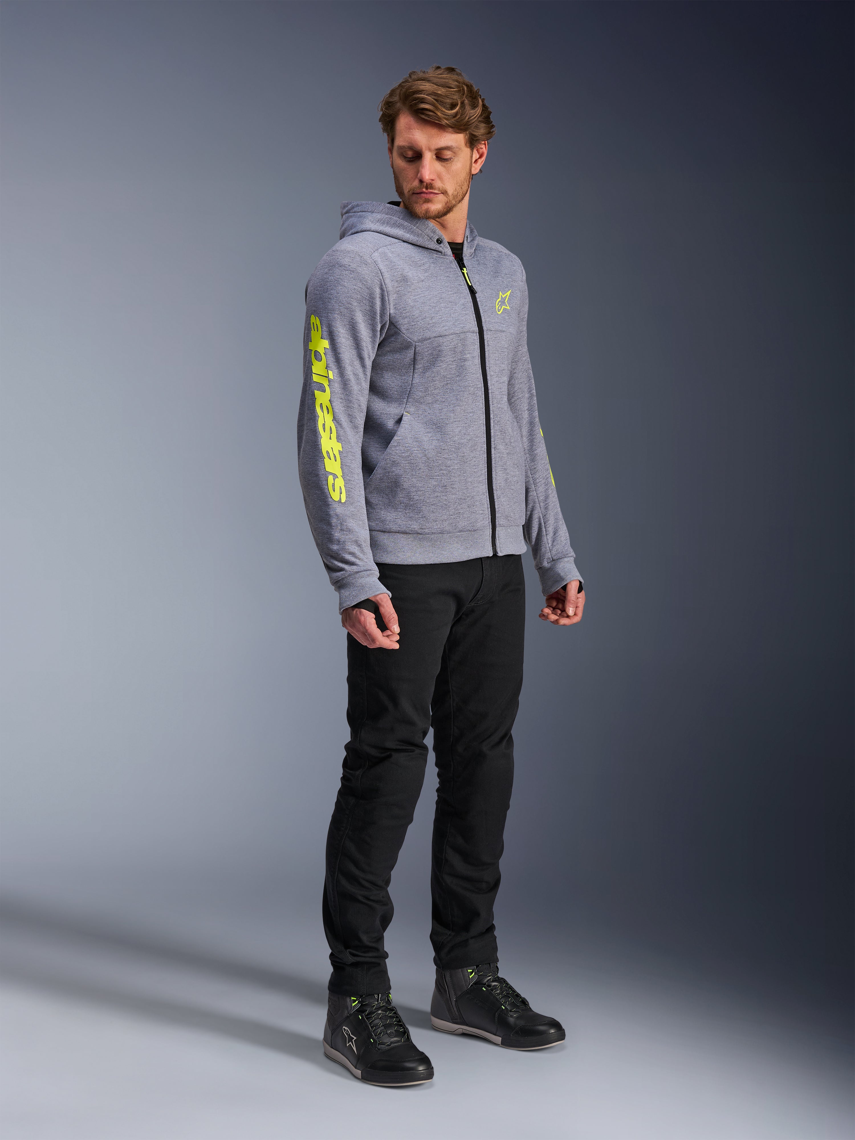 Alpinestars Chrome V2 Sport Hoodie, gråmelerad och fluogul, bärs av en manlig modell med svarta byxor och svarta mc-skor, med framträdande gul logotyp på ärmen och bröstet, avslappnad urban motorcykelutrustning