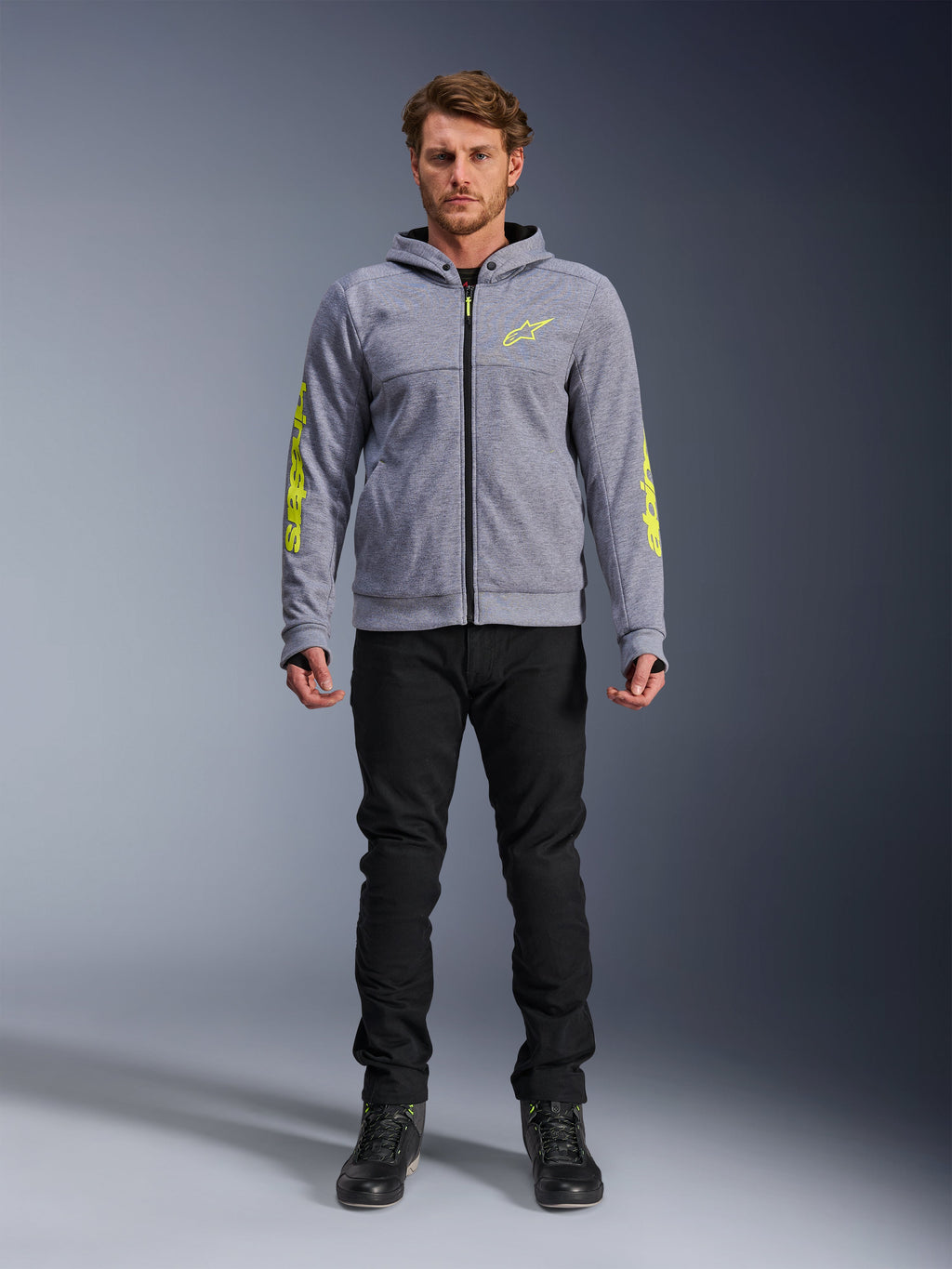 Alpinestars Chrome V2 Sport Hoodie, gråmelerad med fluorescerande gula accenter, urban motorcykelutrustning, visas med svarta byxor och svarta mc-skor, teknisk fleece med tydlig gul logotyp på ärmar och bröst, avslappnad streetstyle för mc-körning