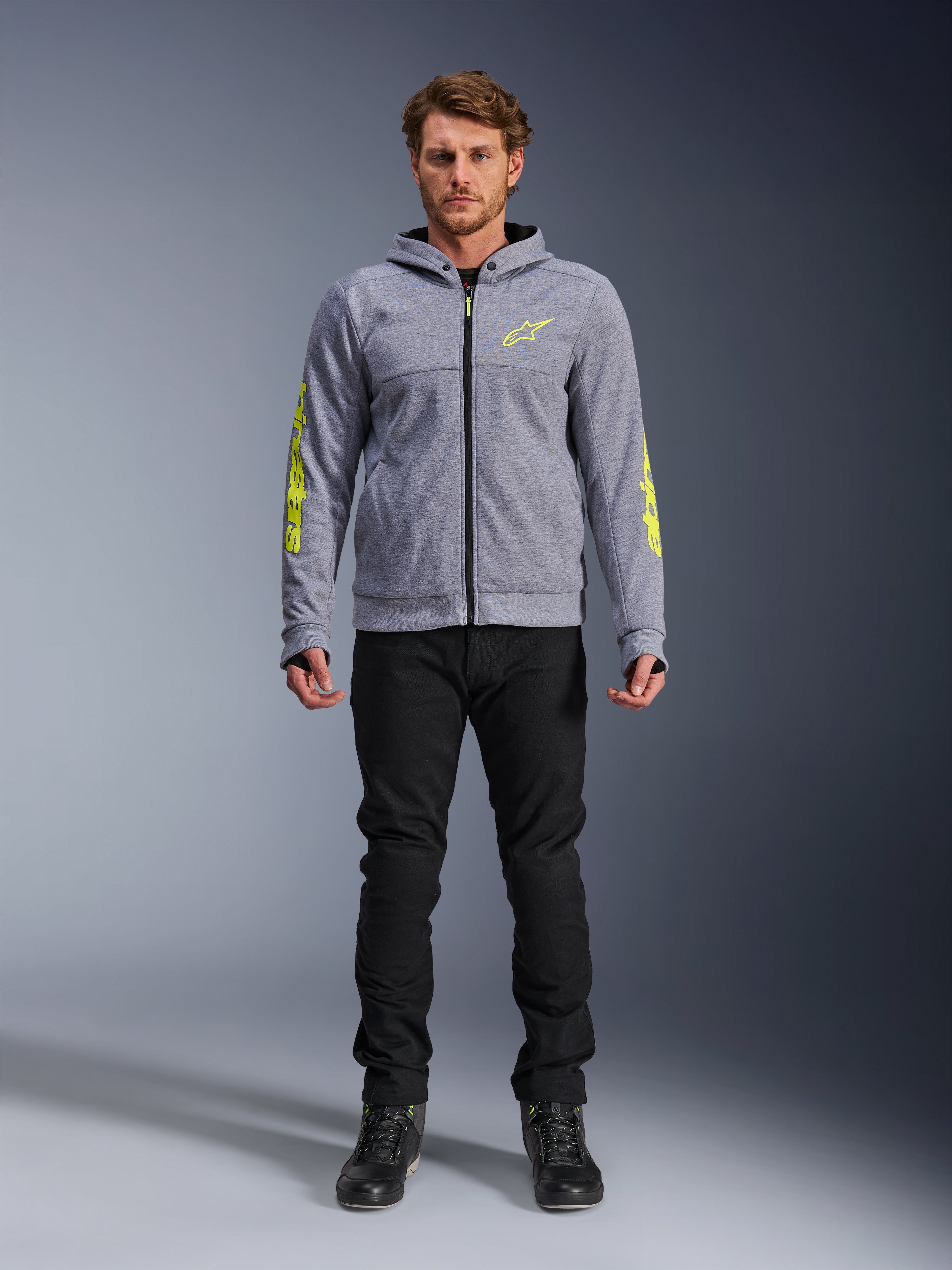 Alpinestars Chrome V2 Sport Hoodie, gråmelerad med fluorescerande gula accenter, urban motorcykelutrustning, visas med svarta byxor och svarta mc-skor, teknisk fleece med tydlig gul logotyp på ärmar och bröst, avslappnad streetstyle för mc-körning