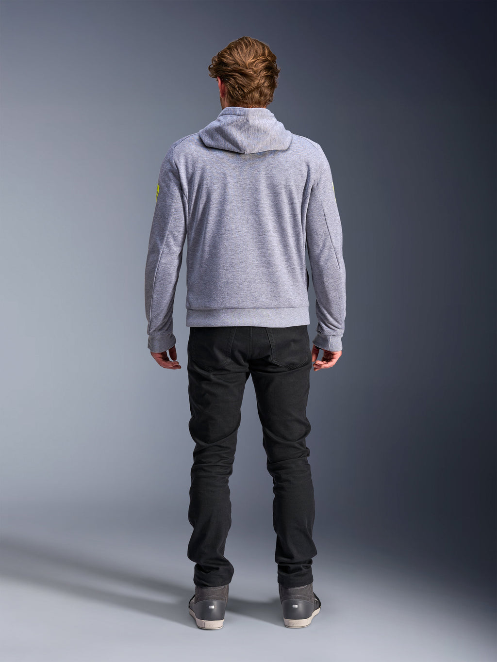 Alpinestars Chrome V2 Sport Hoodie, Gråmelerad Neongul, ryggvy av urban motorcykelutrustning, teknisk huvtröja i fleece buren tillsammans med svarta byxor, med neongula detaljer på ärmarna.