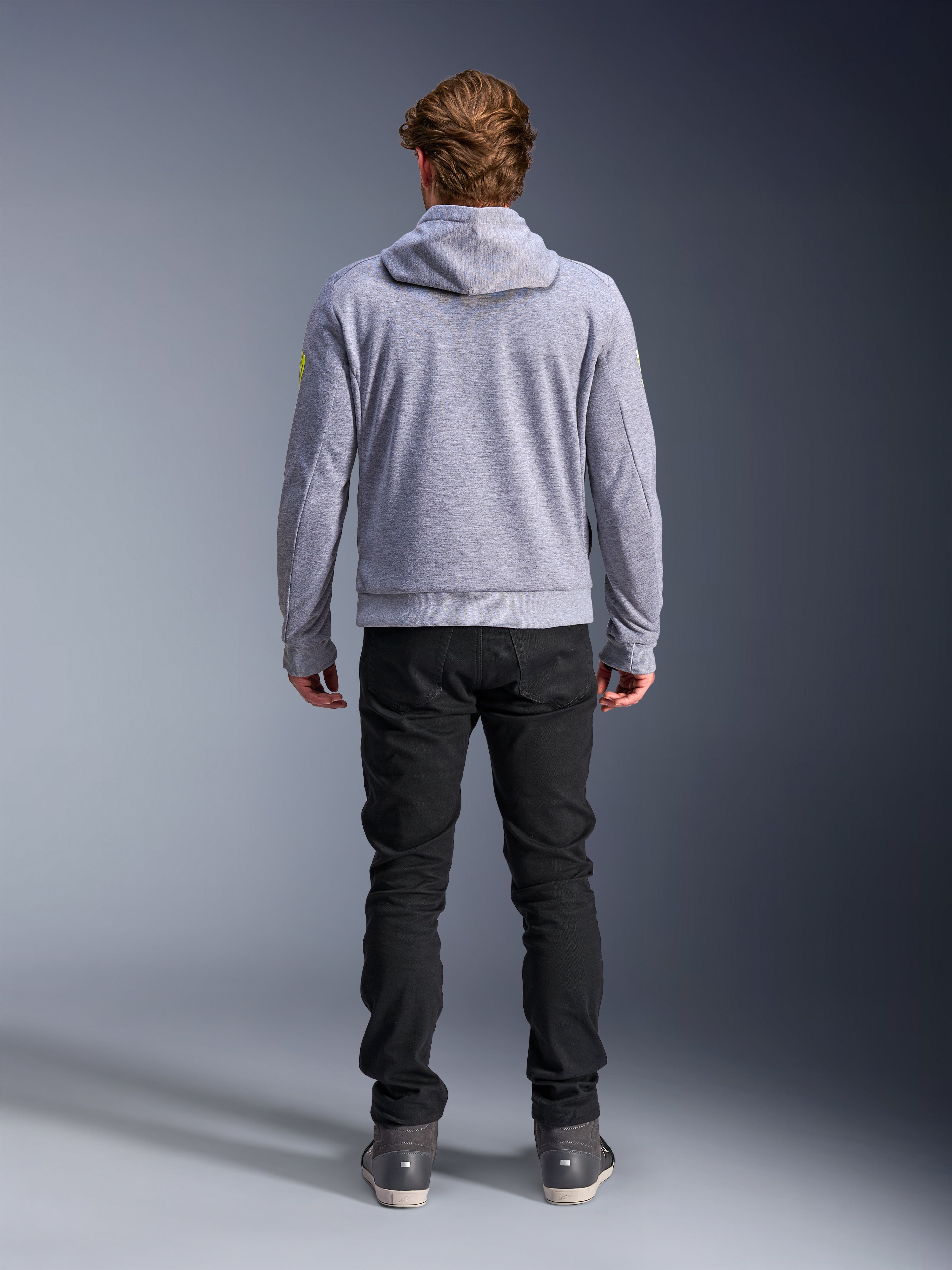 Alpinestars Chrome V2 Sport Hoodie, Gråmelerad Neongul, ryggvy av urban motorcykelutrustning, teknisk huvtröja i fleece buren tillsammans med svarta byxor, med neongula detaljer på ärmarna.