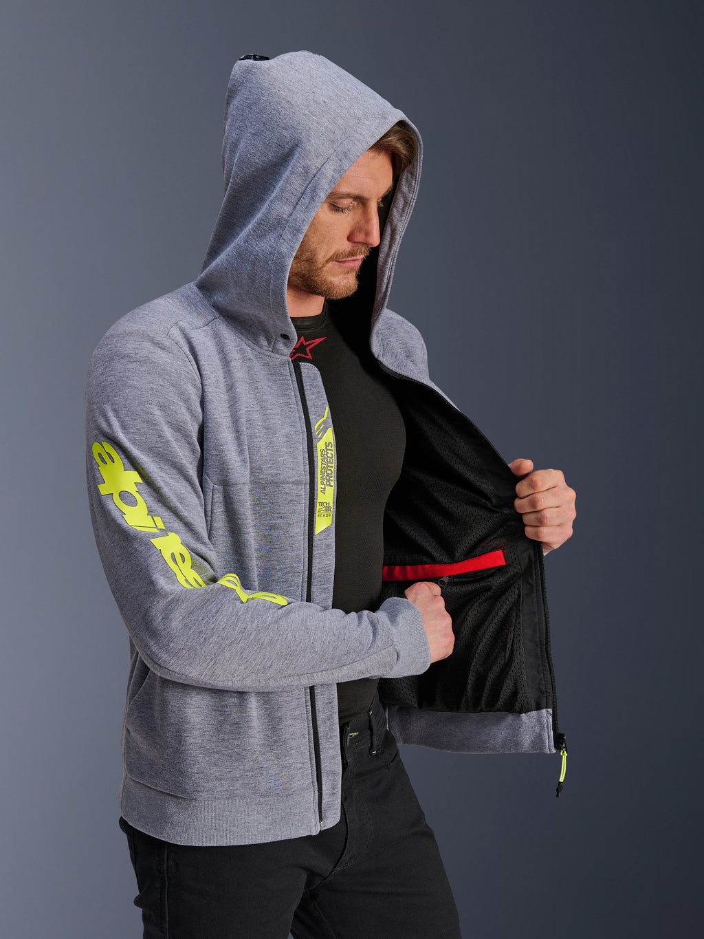 Alpinestars Chrome V2 Sport Hoodie, gråmelerad och fluorescerande gul, man bär huvtröja med huvan uppfälld, öppen dragkedja som visar svart meshfoder och invändig röd dragkedjeförsedd ficka, stor gul logotyp på ärmen, avslappnad urban motorcykelutrustning