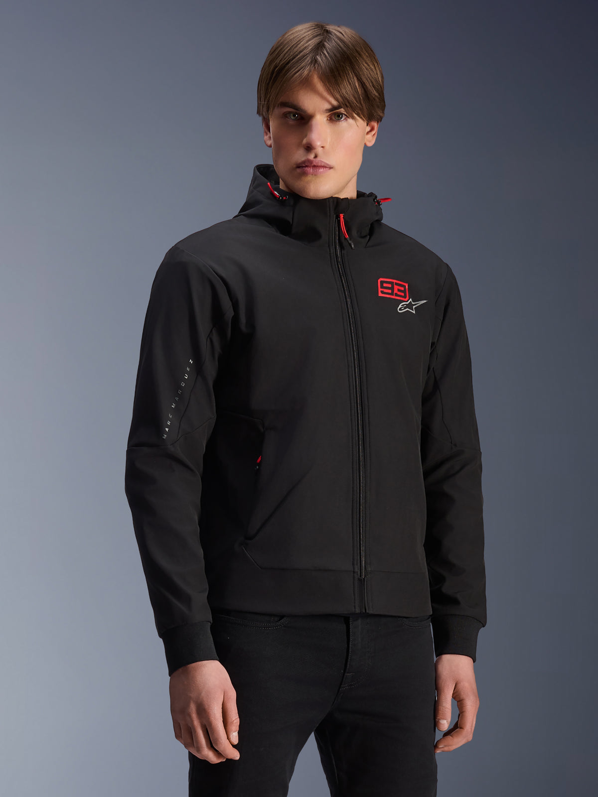 MM93 Radium Tech Hoodie, black urban motorcycle zip-up jacket, red 93 and white Alpinestars logo on chest, worn by model with black pants, featuring a removable hood with red drawstrings and sleeve branding, casual motorsport-inspired design