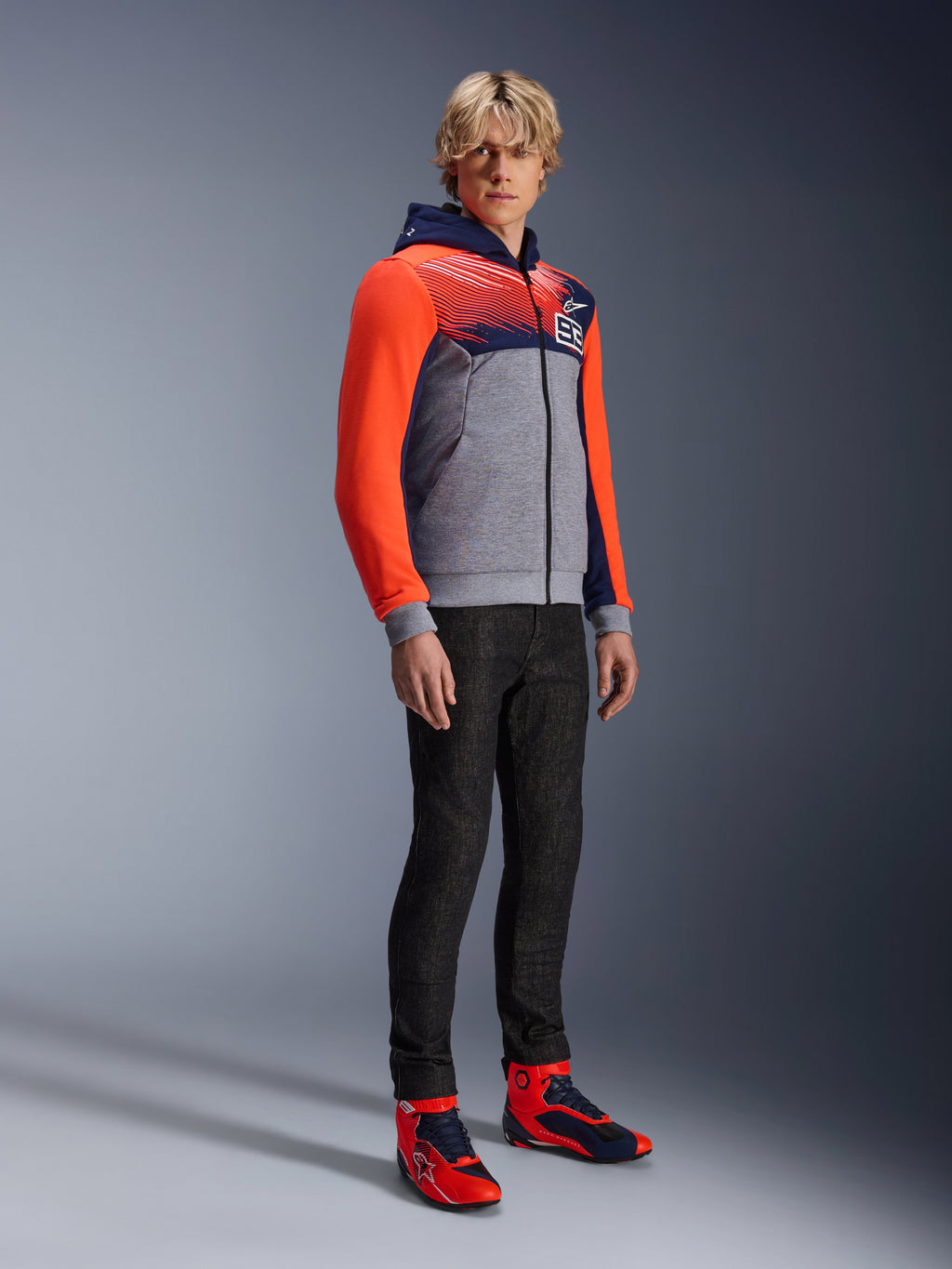 Alpinestars MM93 Chrome V2 Sport Hoodie, urban motorcycle hoodie, gray melange body with navy blue chest and bright red fluo sleeves, featuring the 93 logo on the chest, worn by a model with slim-fit black pants and coordinated red and blue high-top riding shoes, modern athletic fit for street riding