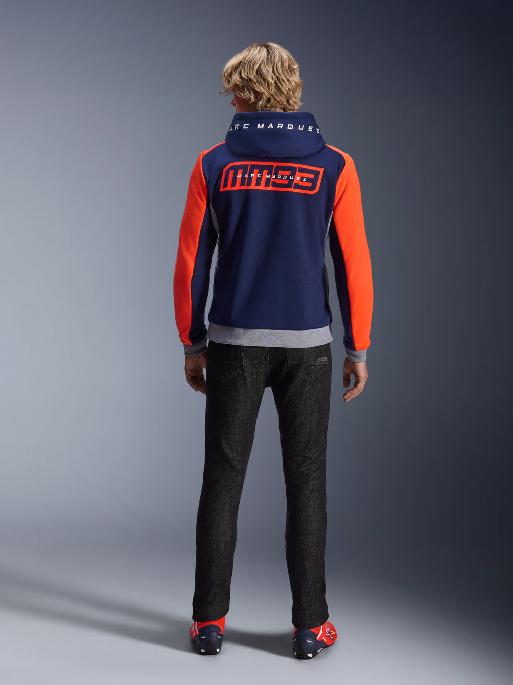Model from the back wearing the Alpinestars MM93 Chrome V2 Sport Hoodie, Blue Navy with Bright Red Fluo sleeves and Gray Mel details, large MM93 and Marc Marquez logos on the back and hood, casual motorcycle riding hoodie paired with black pants