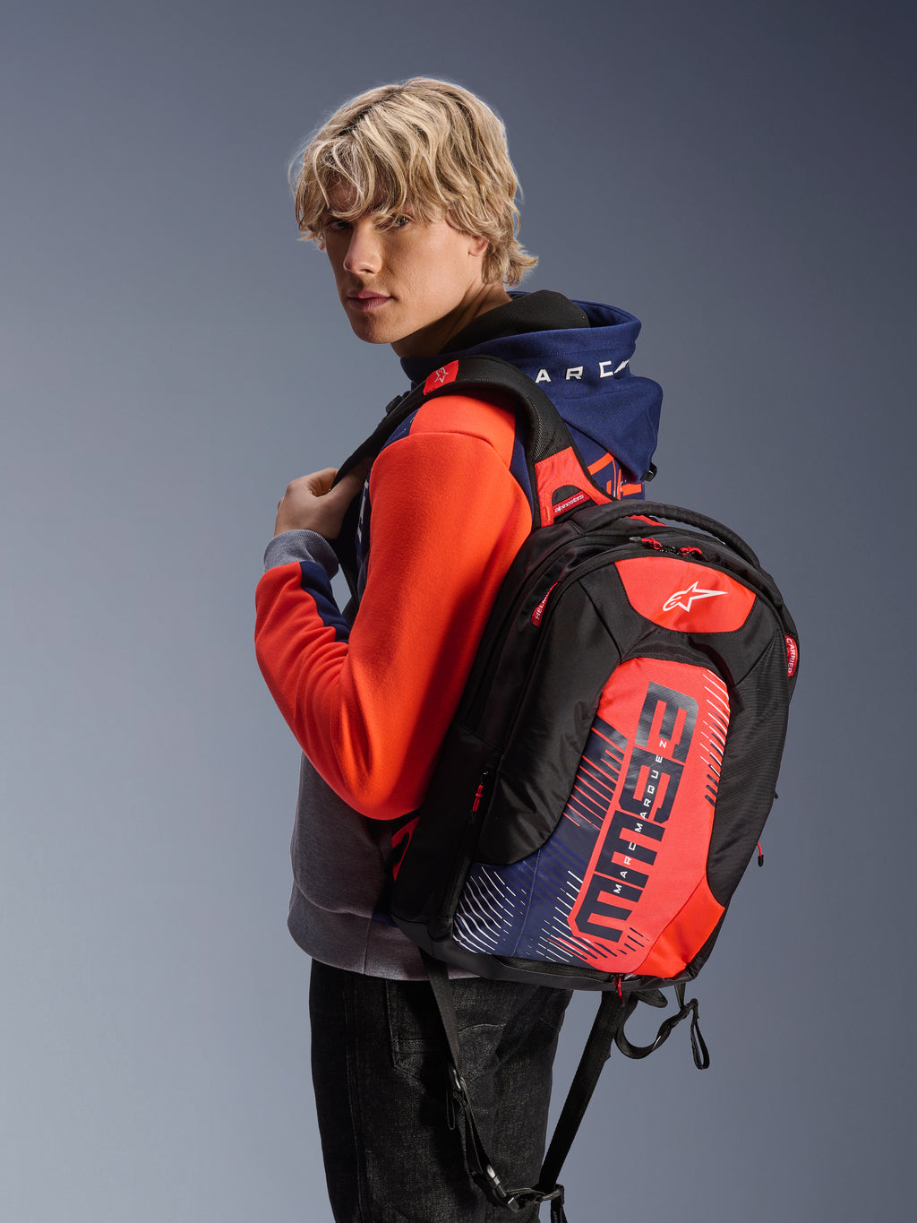 Male model wearing the Alpinestars MM93 Chrome V2 Sport Hoodie in Blue Navy Gray Mel and Red Fluo, paired with a matching MM93 branded backpack, casual urban motorcycle apparel with a modern athletic silhouette.