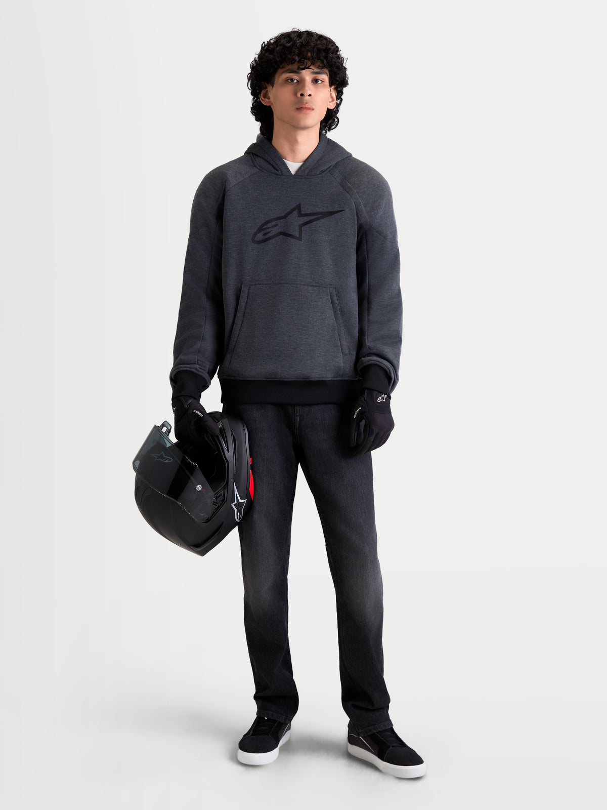 Alpinestars Ageless jacka med huva, urban motorcykeljacka för herr i mörkgrå melange, framifrån på en modell som bär svarta MC-handskar och håller en hjälm, med integrerat skydd på axlar och armbågar
