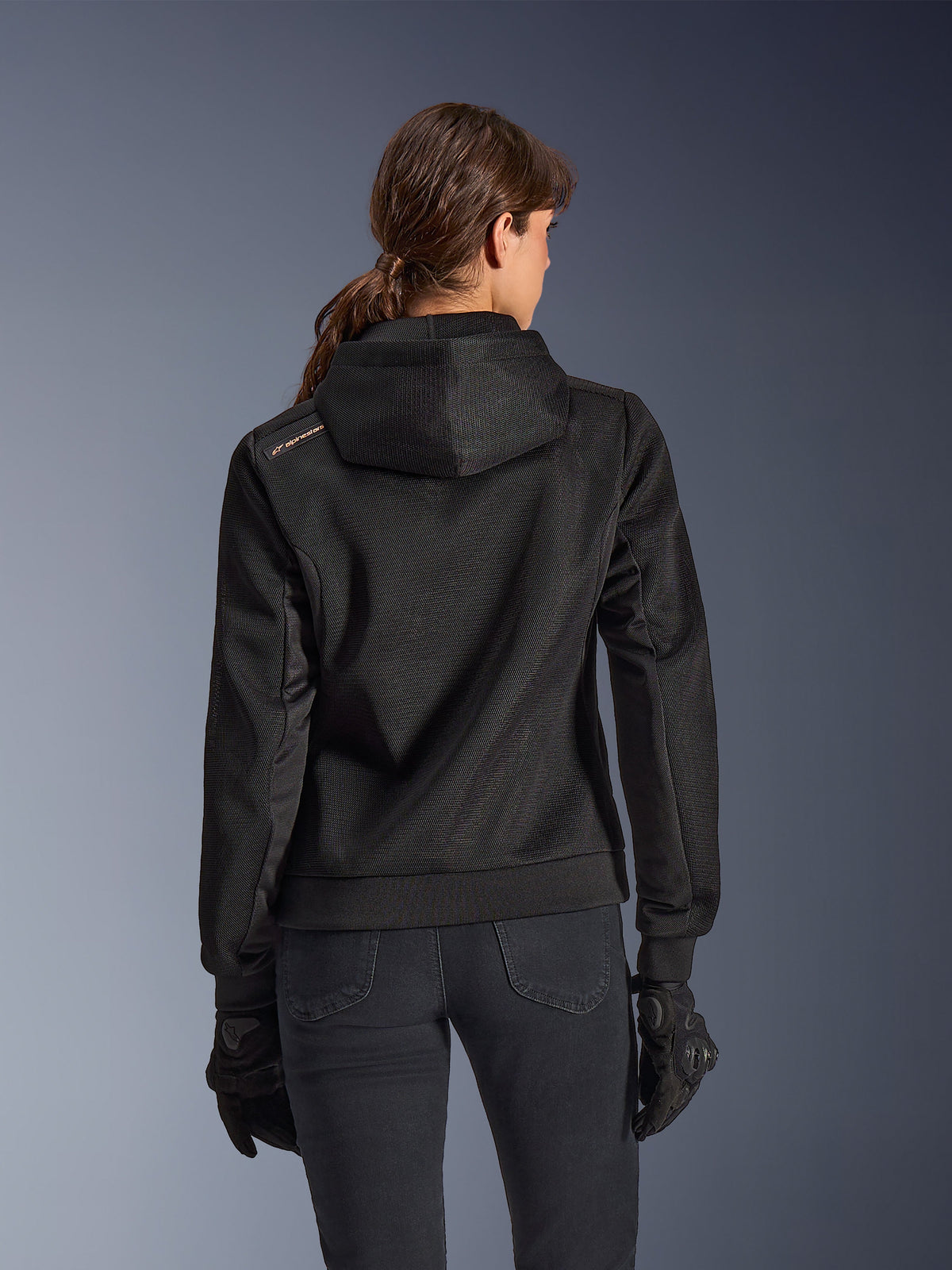 Alpinestars Stella Chrome Superairflow Hoodie, vy bakifrån av kvinna som bär svart teknisk motorcykelhoodie i 3D-mesh, har en konstruerad huva och kopparfärgad logotyp på axeln, bärs med svarta motorcykelhandskar och byxor.