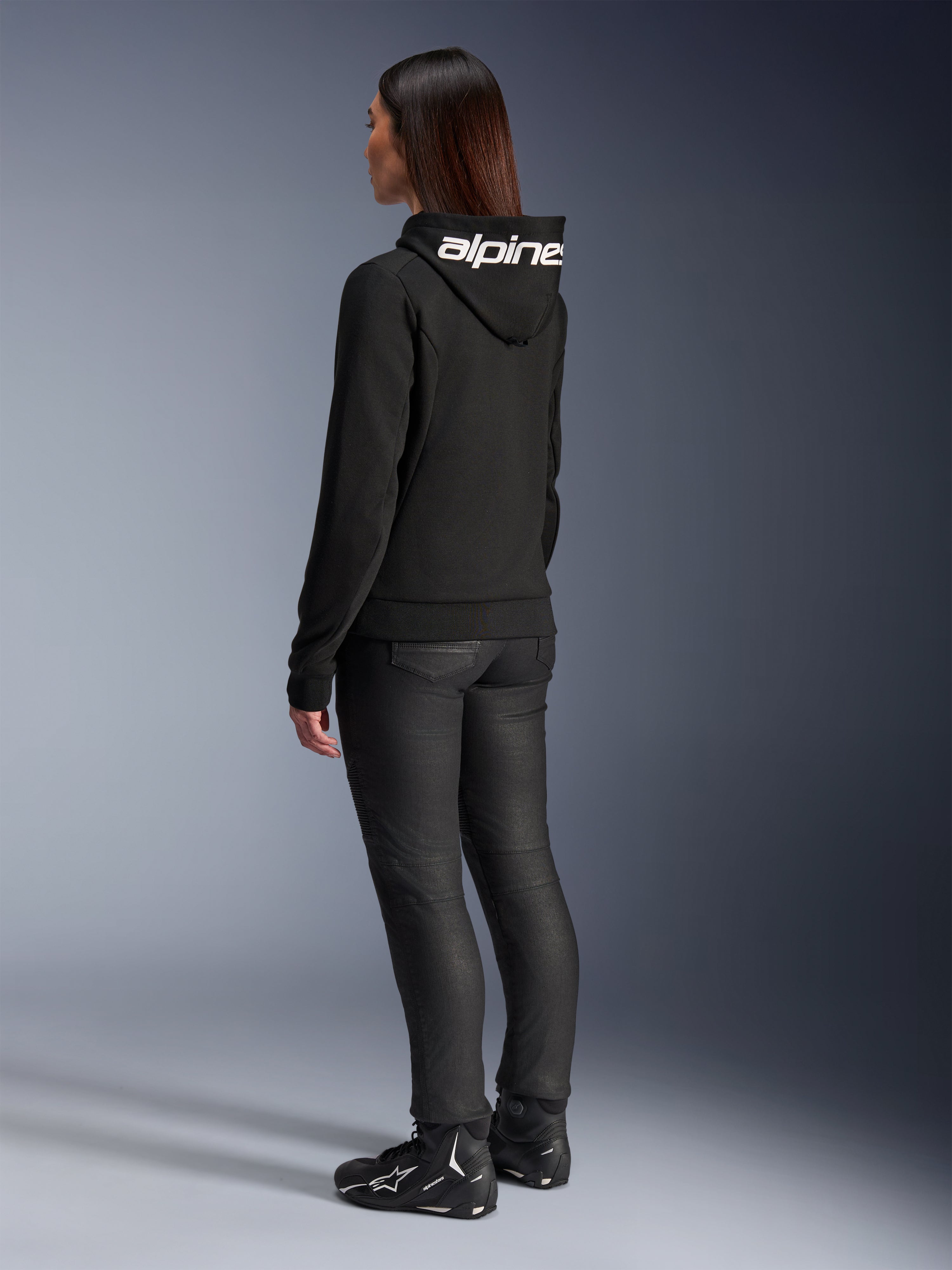 Woman wearing Alpinestars Stella Chrome V2 Sport Hoodie, rear view, black motorcycle hoodie with white Alpinestars logo on the hood, paired with black technical riding pants and black motorcycle shoes, casual urban commuting gear