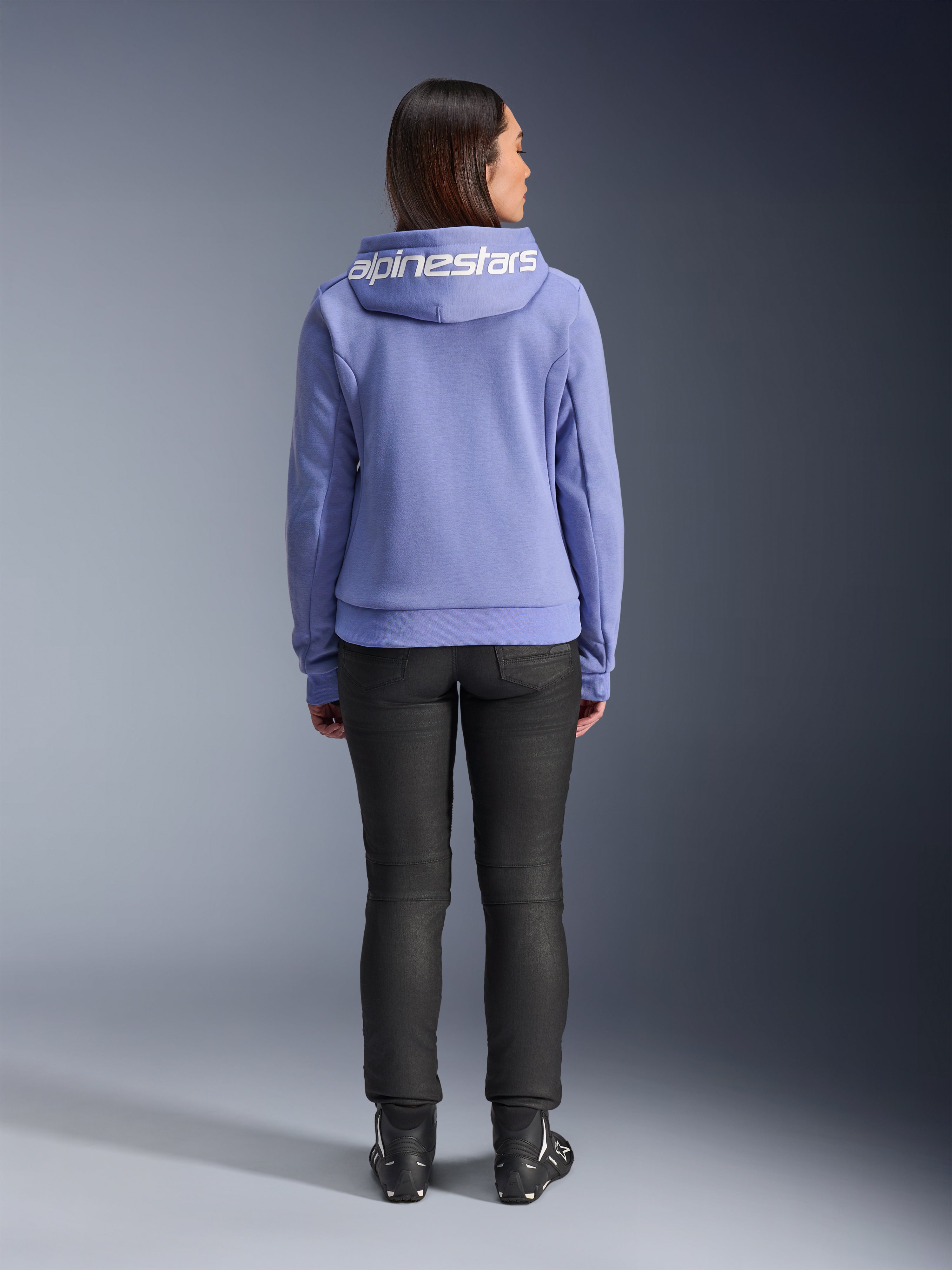 Alpinestars Stella Chrome V2 Sport Hoodie, Blue Violet, ryggvy av en kvinna som bär huvtröjan med vit Alpinestars-logotyp över huvan, kombinerad med svarta byxor och MC-stövlar, urban MC-utrustning i vardaglig stil