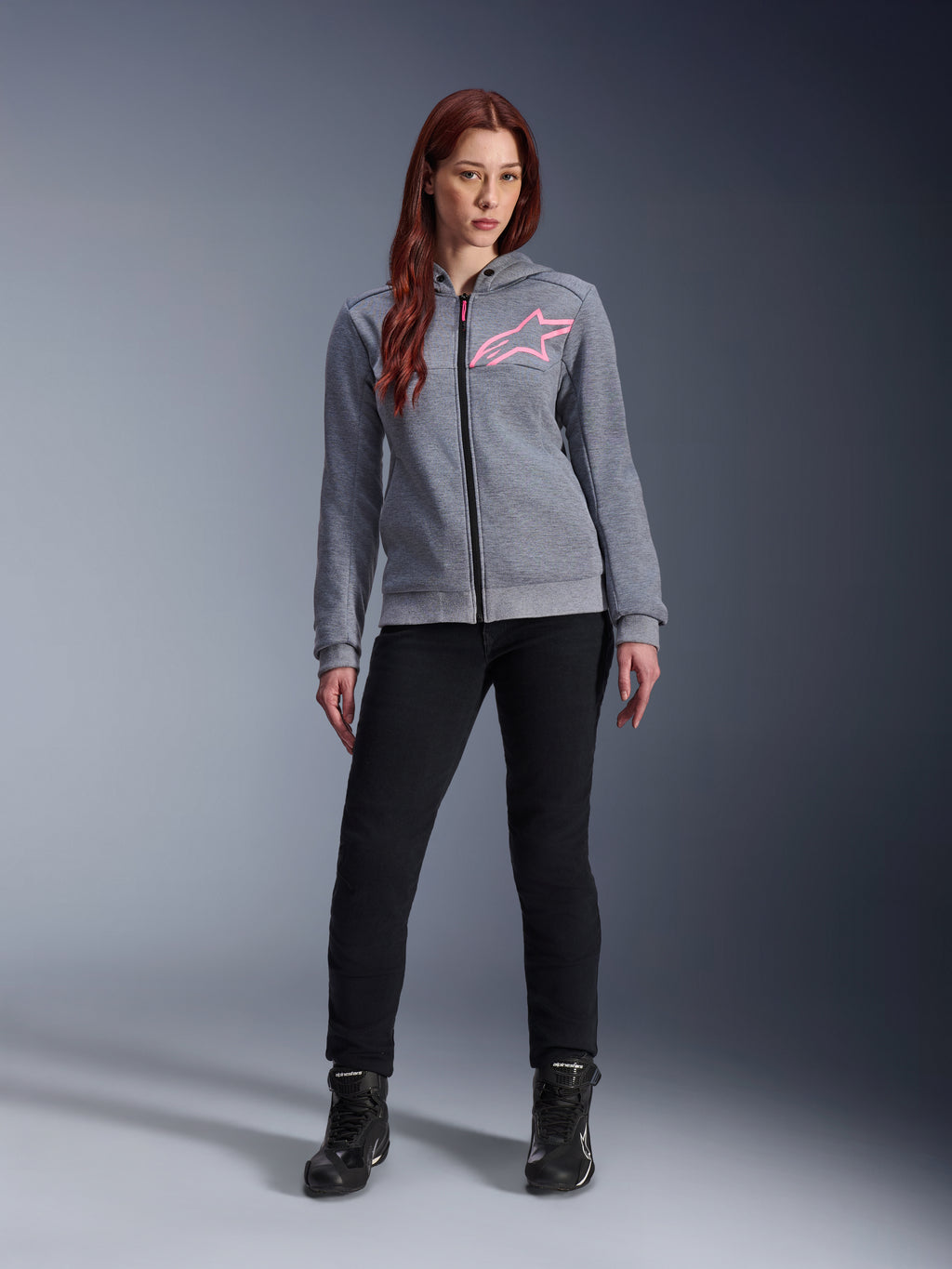 Alpinestars Stella Chrome V2 Sport Hoodie, gråmelerad och fluorescerande rosa, bärs av en kvinna med svarta byxor och stövlar, med en rosa Alpinestars-stjärnlogotyp på bröstet, urban motorcykelutrustning med hel dragkedja, lätt teknisk fleece med en avslappnad atletisk passform.