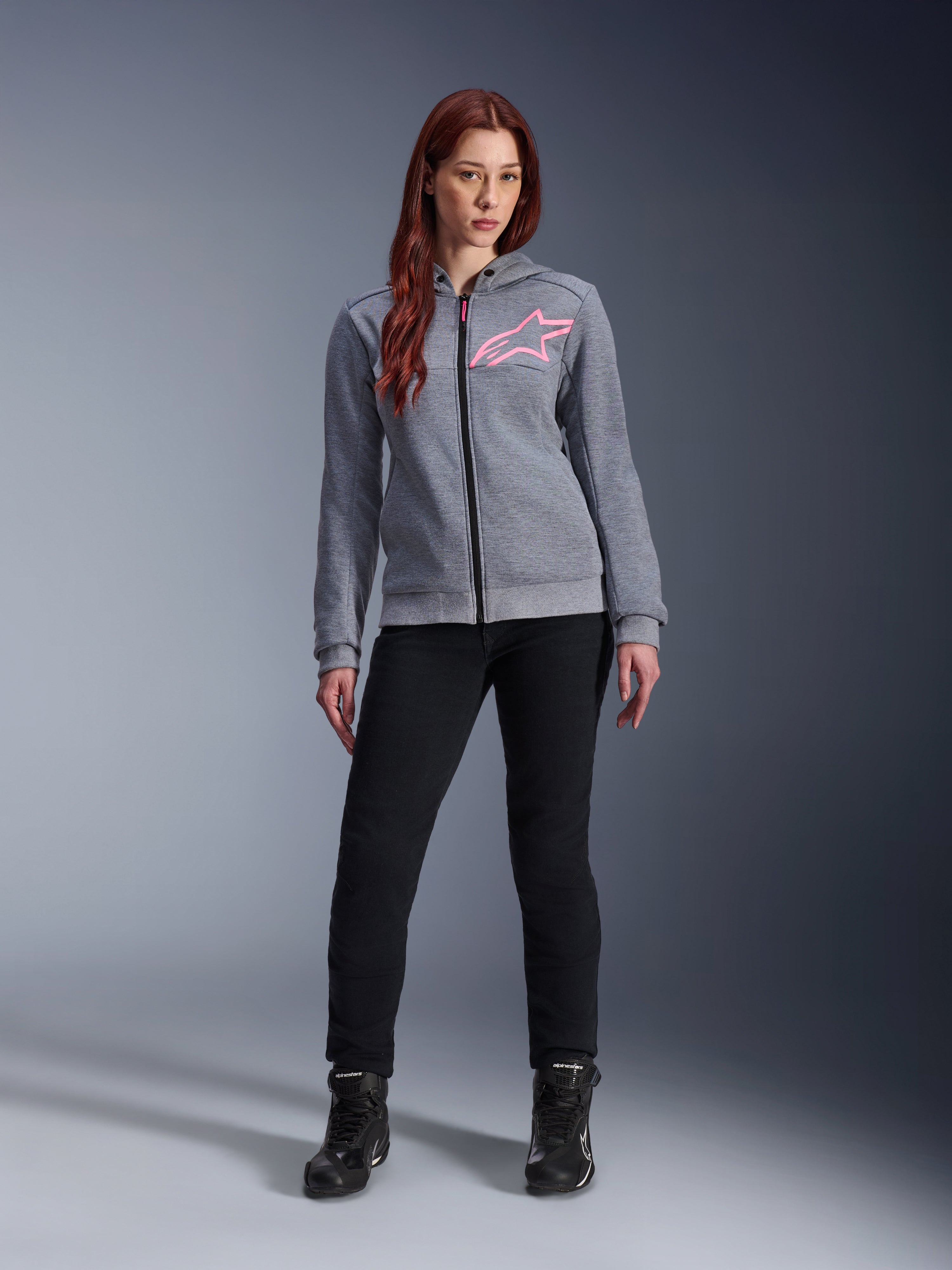 Alpinestars Stella Chrome V2 Sport Hoodie, gråmelerad och fluorescerande rosa, bärs av en kvinna med svarta byxor och stövlar, med en rosa Alpinestars-stjärnlogotyp på bröstet, urban motorcykelutrustning med hel dragkedja, lätt teknisk fleece med en avslappnad atletisk passform.