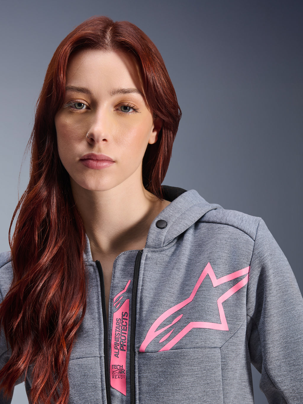 Kvinna i Alpinestars Stella Chrome V2 Sport Hoodie, Gray Heather Pink Fluo, närbild som visar den rosa Alpinestars-stjärnlogotypen och Tech-Air Ready-märkning, urban motorcykelklädsel.
