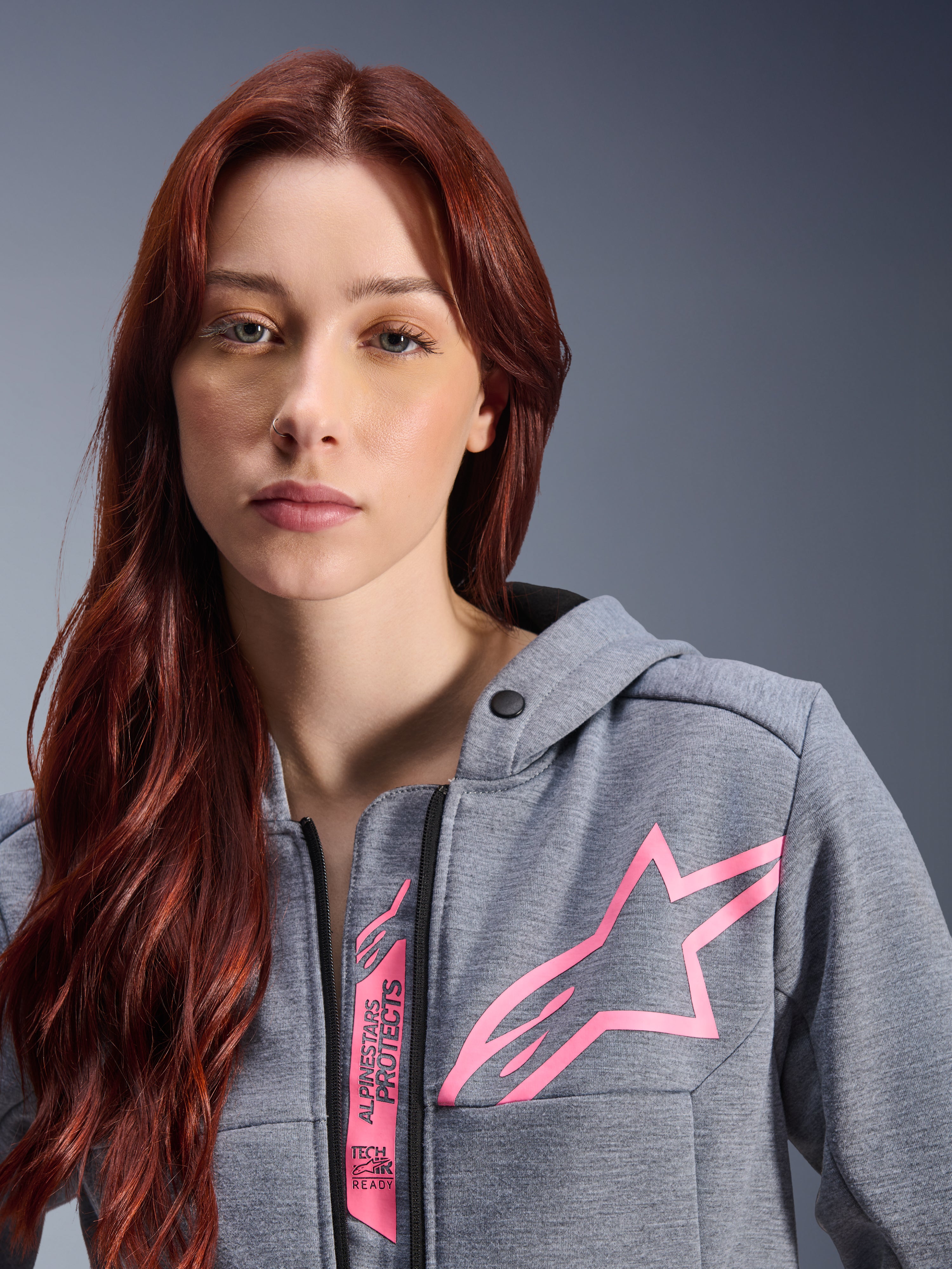 Kvinna i Alpinestars Stella Chrome V2 Sport Hoodie, Gray Heather Pink Fluo, närbild som visar den rosa Alpinestars-stjärnlogotypen och Tech-Air Ready-märkning, urban motorcykelklädsel.