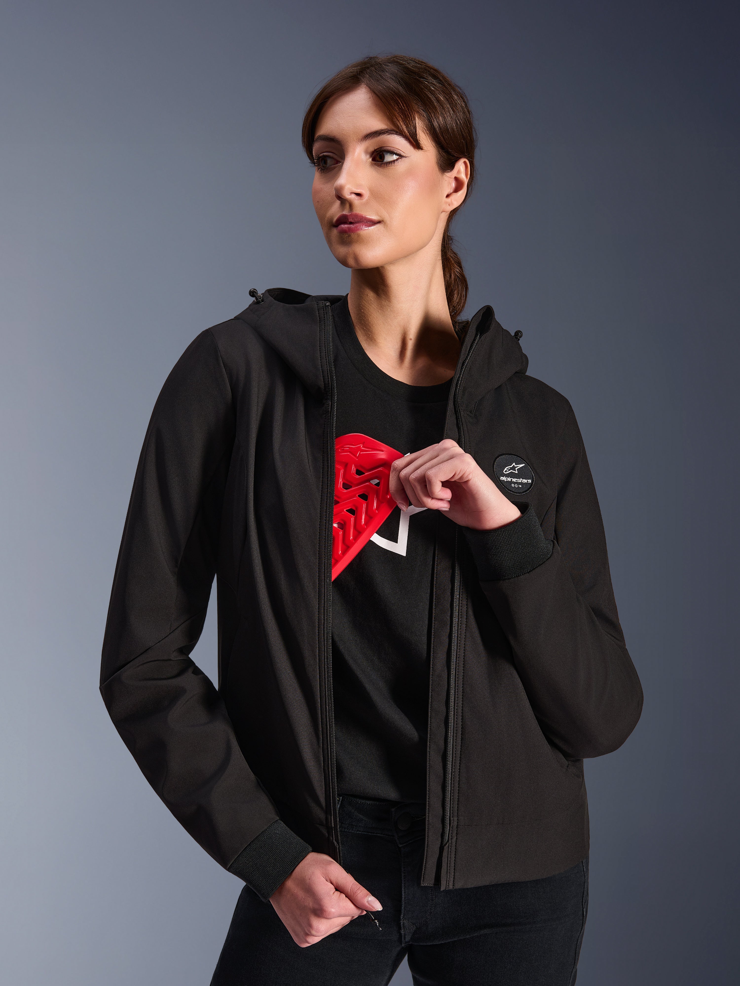 Alpinestars Woman Stella Radium Tech Hoodie, svart huvtröja för stadskörning, kvinnlig modell som visar rött Nucleon Flex Plus-skydd på insidan, lätt 3-lagers vindtätt tekniskt skal med ribbade muddar och logotypmärke på bröstet, skyddande motorcykelutrustning