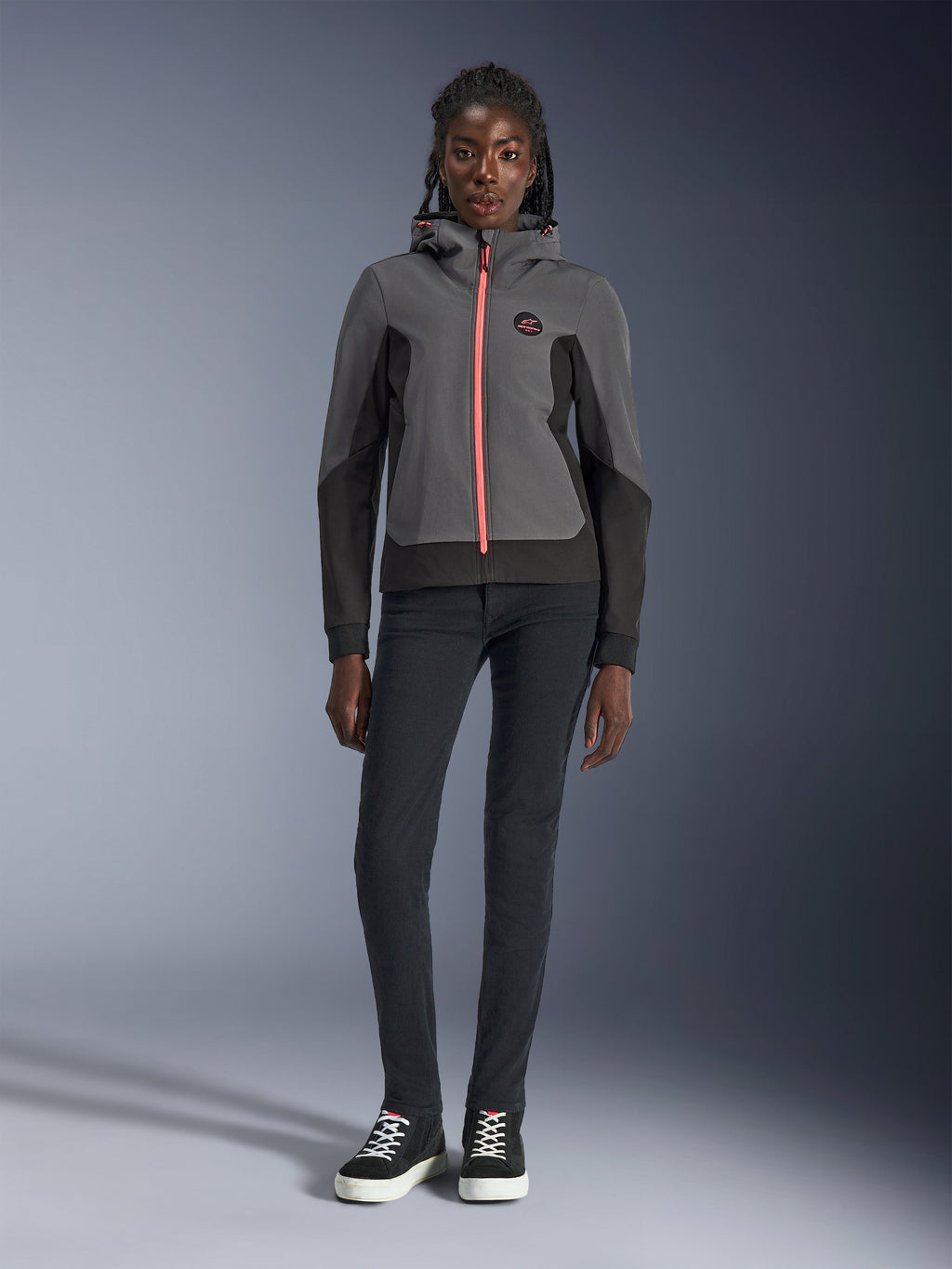Kvinnlig modell bär Alpinestars Stella Radium Tech Hoodie, mörkgrå och svart med en klarorange dragkedja som accent, urban motorcykelhuvtröja matchad med svarta slim-fit-byxor och sneakers, avslappnad atletisk prestandautrustning för damer.