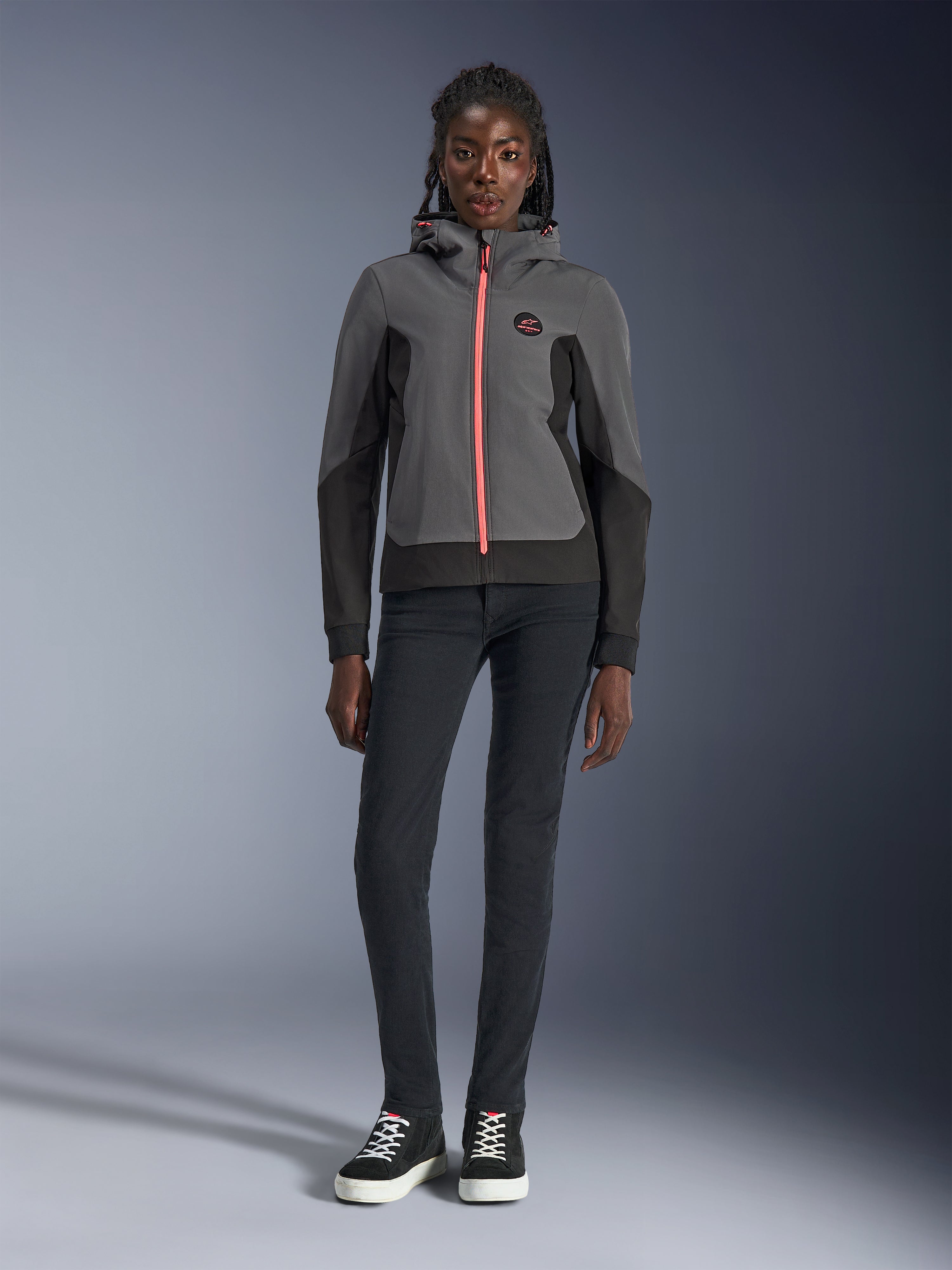 Kvinnlig modell bär Alpinestars Stella Radium Tech Hoodie, mörkgrå och svart med en klarorange dragkedja som accent, urban motorcykelhuvtröja matchad med svarta slim-fit-byxor och sneakers, avslappnad atletisk prestandautrustning för damer.