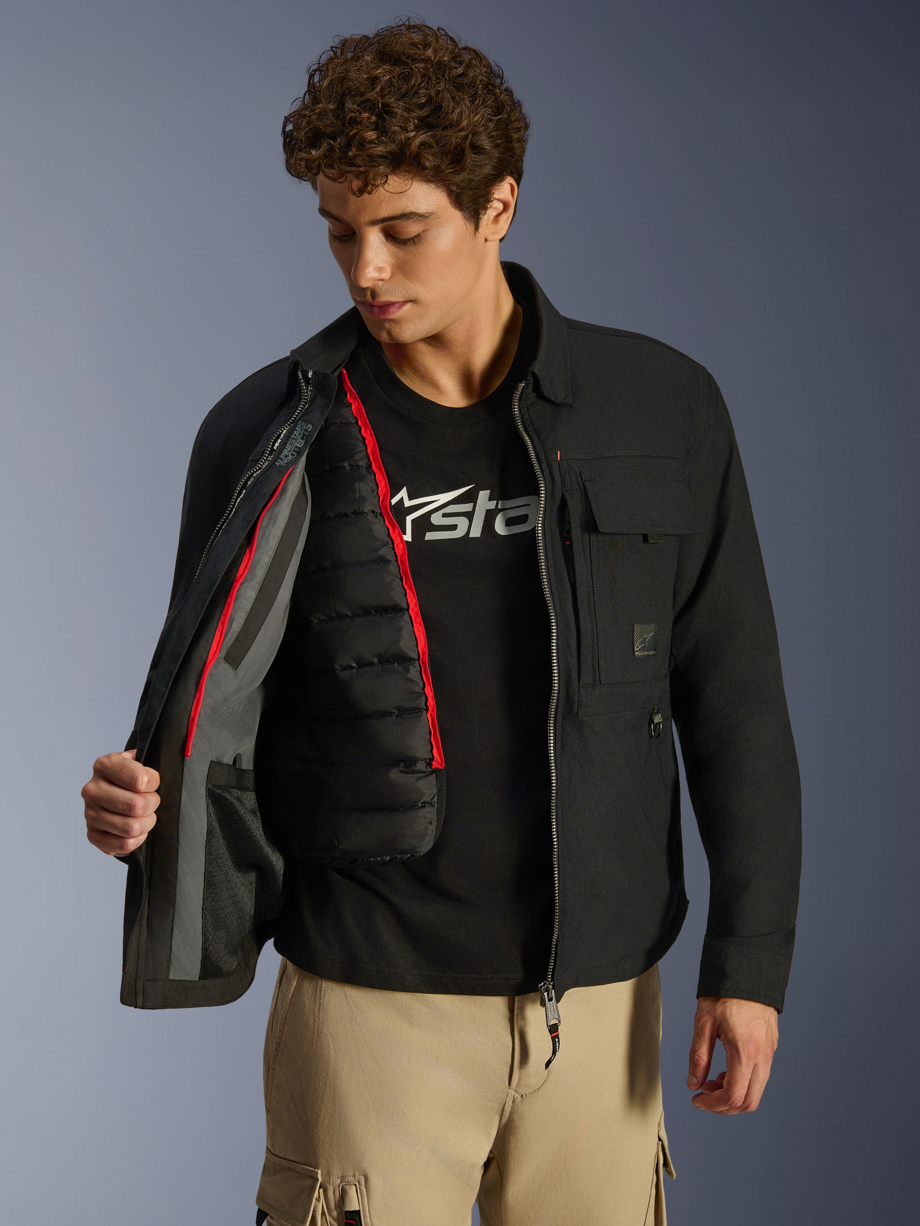 Alpinestars Bryon WR överskjorta, svart, urban motorcykeljacka som visas öppen för att visa en avtagbar svart kviltad termoväst med röda detaljer, bärs med beige cargobyxor och en t-shirt med logotyp, funktionell design inspirerad av arbetskläder