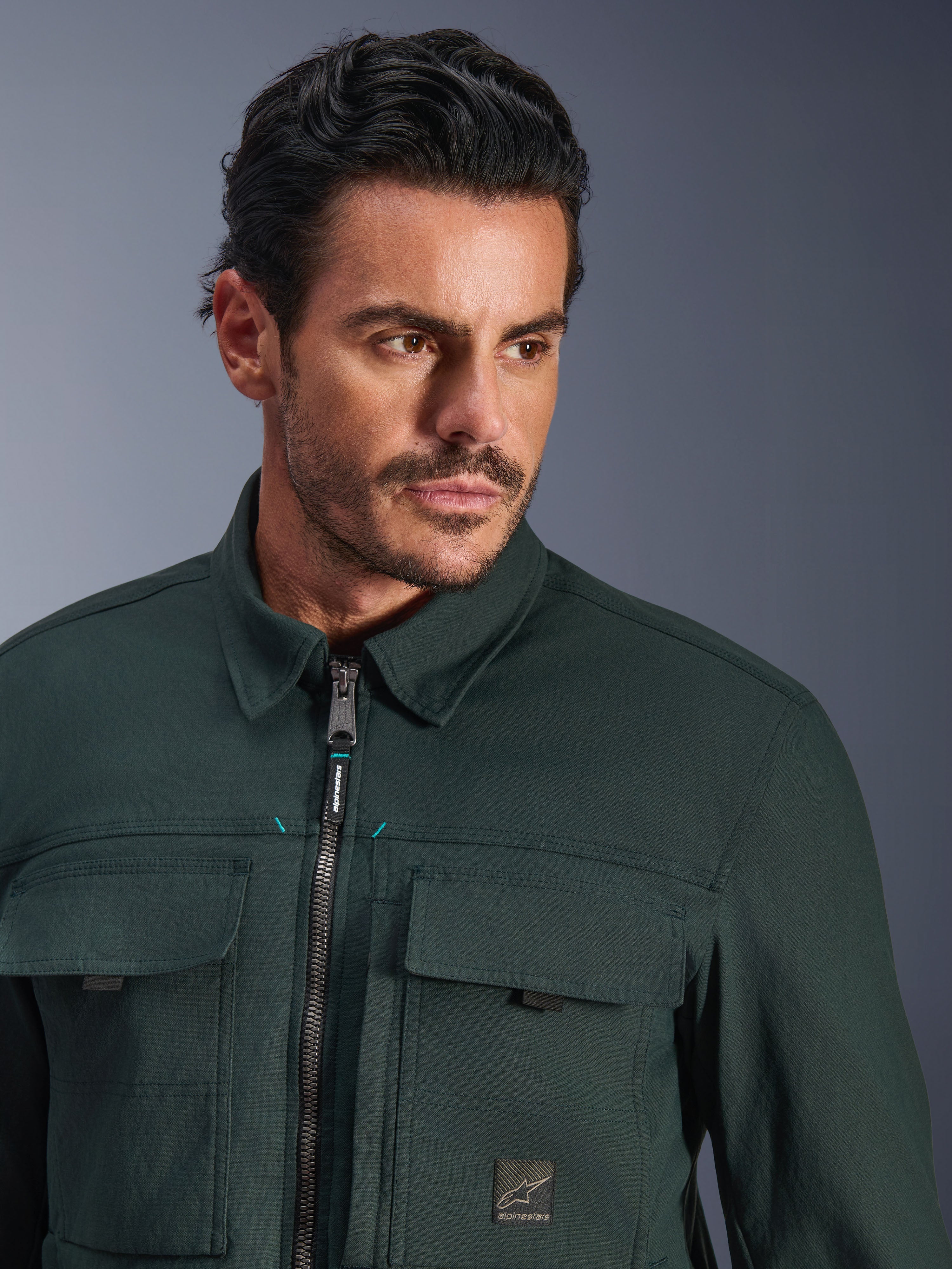 Alpinestars Bryon Water Resistant Overshirt in Dark Emerald, close-up portrait of a man wearing the urban motorcycle jacket, featuring waxed canvas texture, front zipper with subtle teal accents, chest cargo pockets, and branded logo patch.