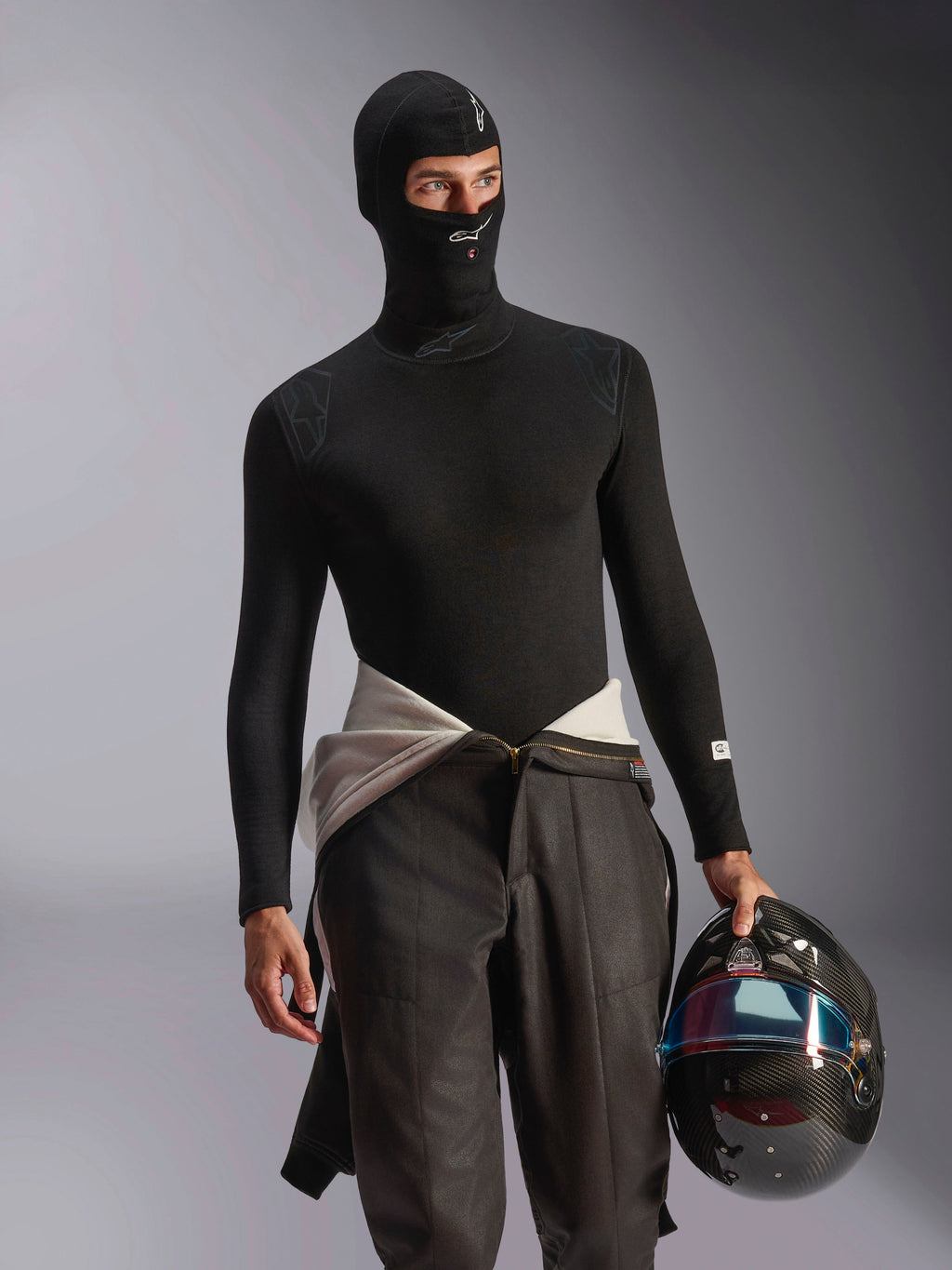 Manlig modell iklädd Alpinestars ZX Evo V3 Balaklava i svart, med vita logotyper på pannan och hakan, kombinerad med en svart långärmad underställströja och hållandes en racinghjälm i kolfiber, professionell flamsäker motorsportutrustning för bilracing.