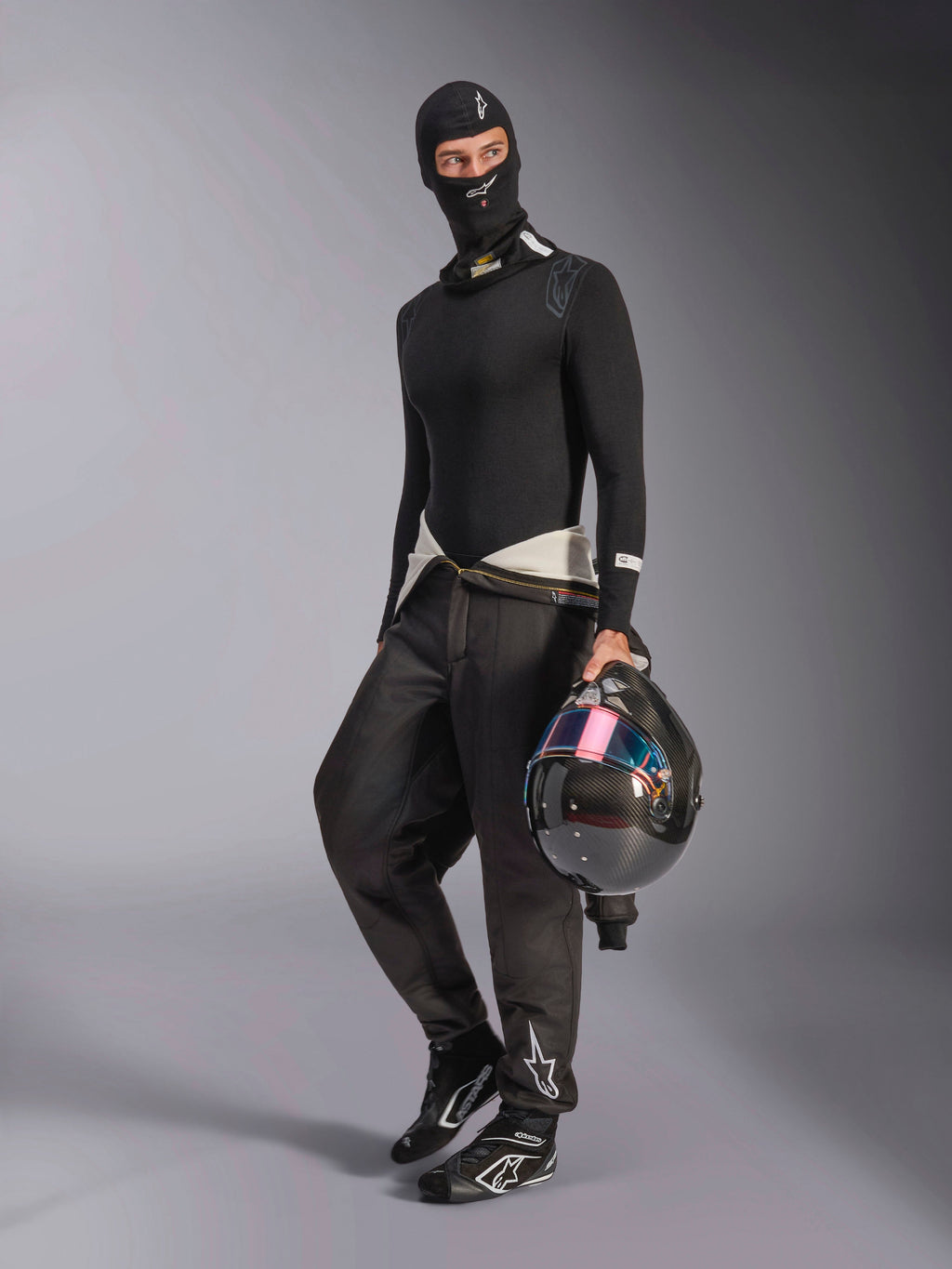 Alpinestars ZX Evo V3 Balaclava, svart, bärs av en förare med komplett underställ för racing, vit logotyp i pannan, föraren håller en kolfiberhjälm med iriserande visir, professionella flamsäkra motorsportkläder.