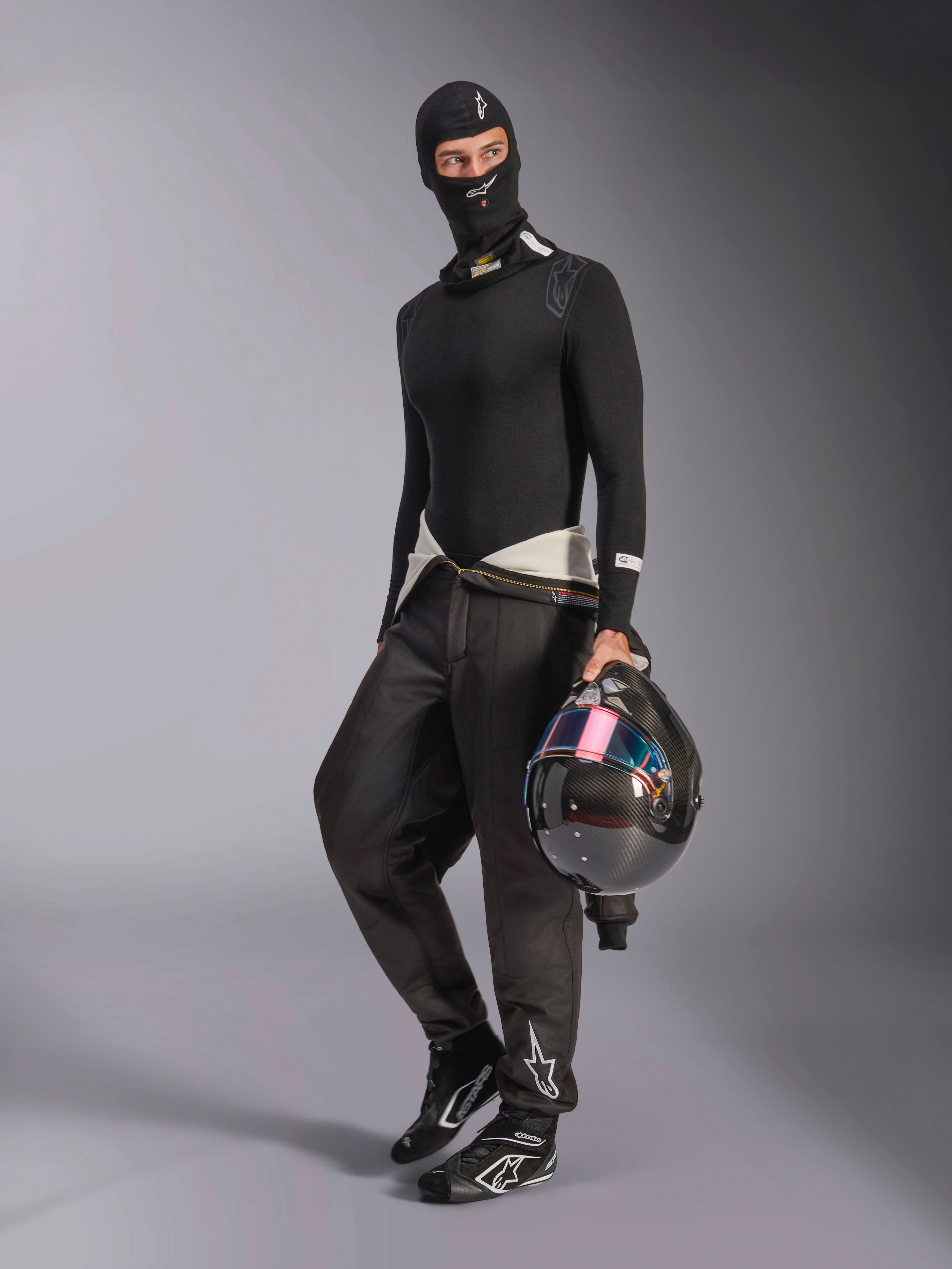 Alpinestars ZX Evo V3 Balaclava, svart, bärs av en förare med komplett underställ för racing, vit logotyp i pannan, föraren håller en kolfiberhjälm med iriserande visir, professionella flamsäkra motorsportkläder.