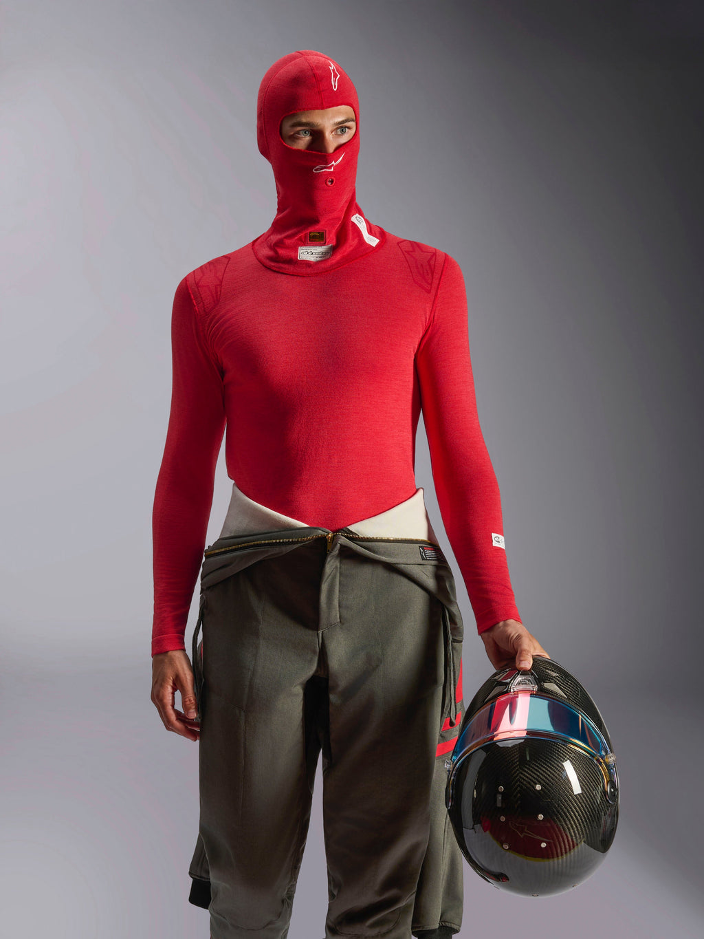 Alpinestars ZX Evo V3 Balaklava i rött, underställ för bilsport bärs av förare med matchande röd långärmad tröja och olivgröna racingbyxor, håller en kolfiberhjälm, professionell flamsäker motorsportutrustning