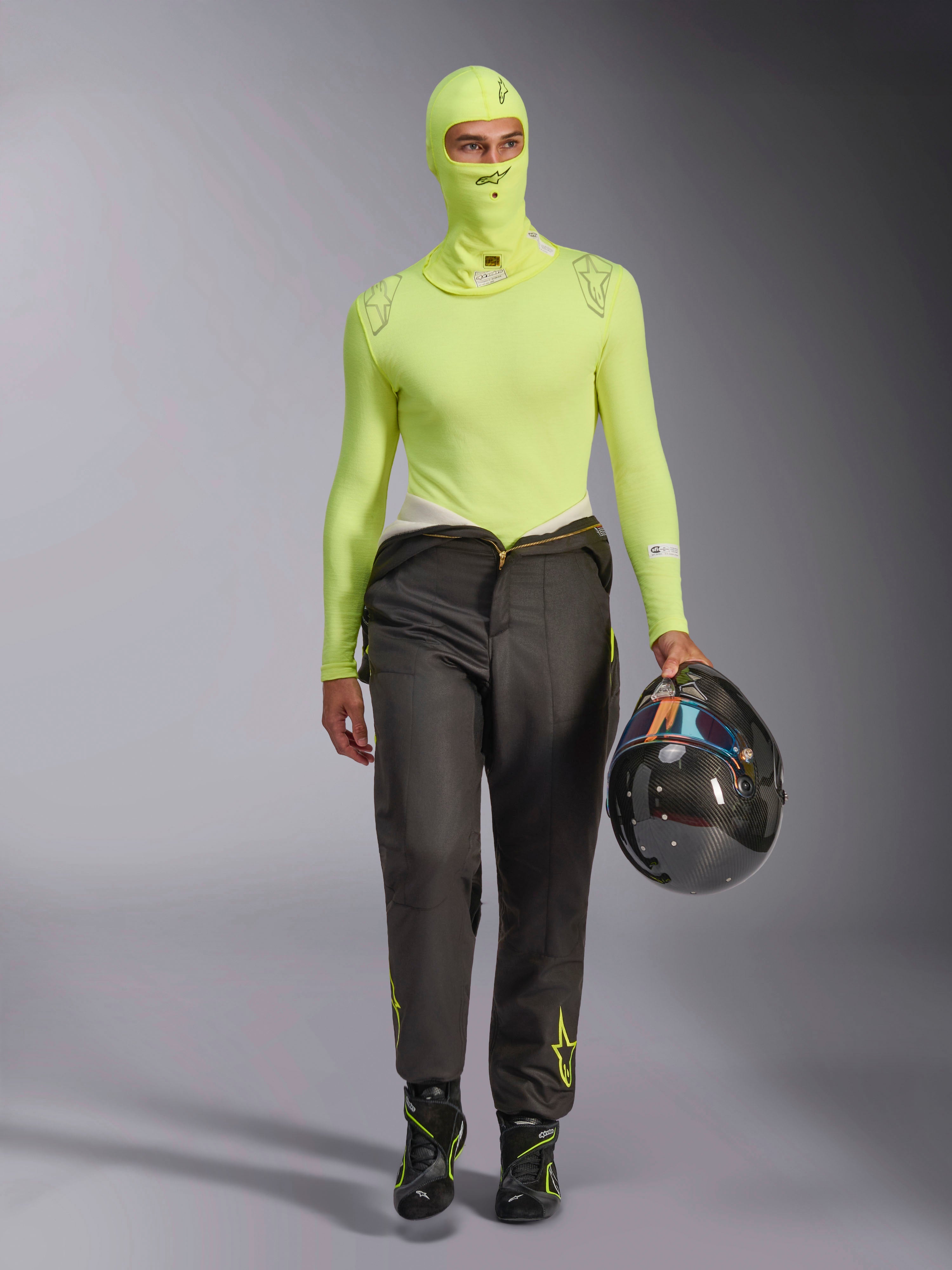 Alpinestars ZX Evo V3 Balaklava i Yellow Fluo, buren med matchande långärmad underställströja och mörkgrå racingbyxor, manlig modell som håller en kolfiberhjälm, professionell racingutrustning