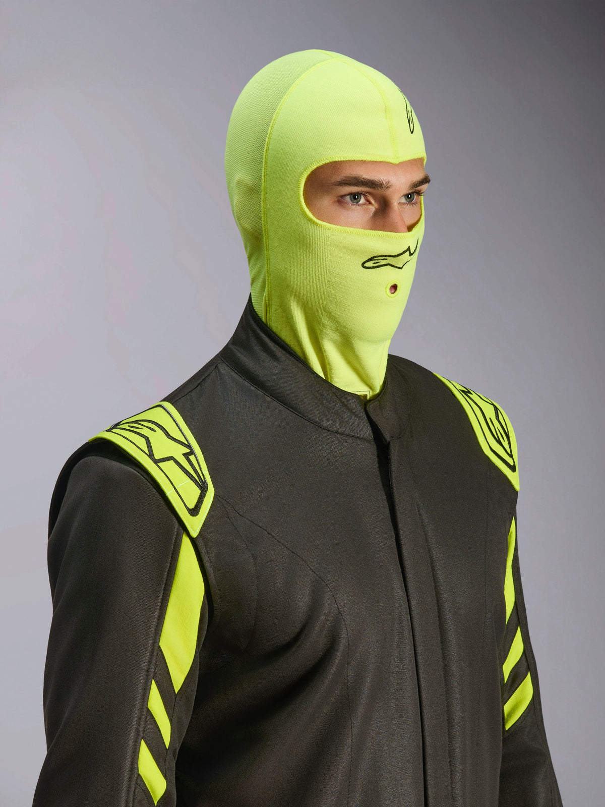 Manlig modell som bär Alpinestars ZX Evo V3 Balaclava i Yellow Fluo, flamsäker racing-balaklava, kombinerad med en mörkgrå racingoverall med neongula detaljer, med en svart Alpinestars-logotyp och munöppning för hydratiseringsslang, professionell skyddsutrustning för motorsport