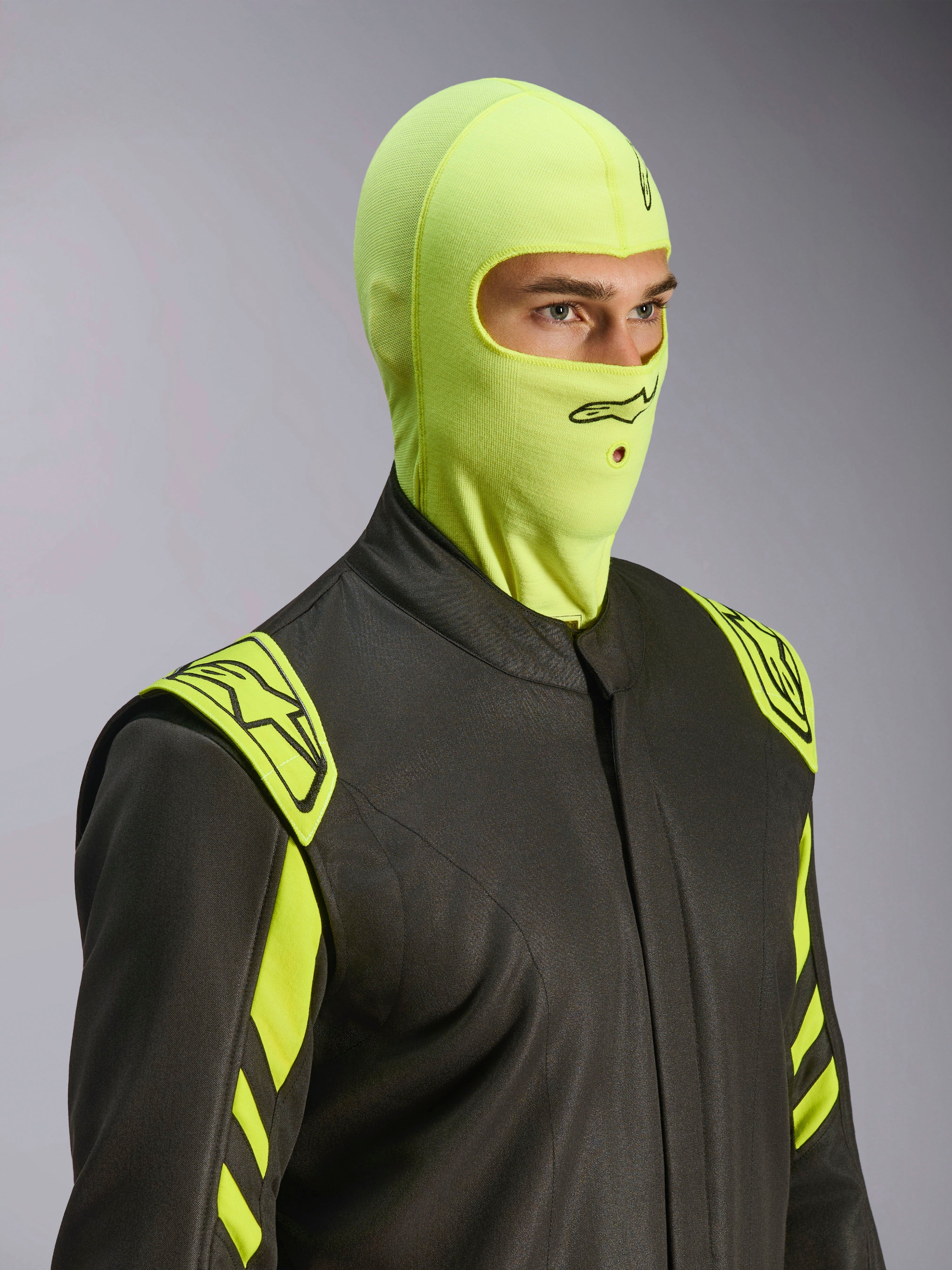 Manlig modell som bär Alpinestars ZX Evo V3 Balaclava i Yellow Fluo, flamsäker racing-balaklava, kombinerad med en mörkgrå racingoverall med neongula detaljer, med en svart Alpinestars-logotyp och munöppning för hydratiseringsslang, professionell skyddsutrustning för motorsport