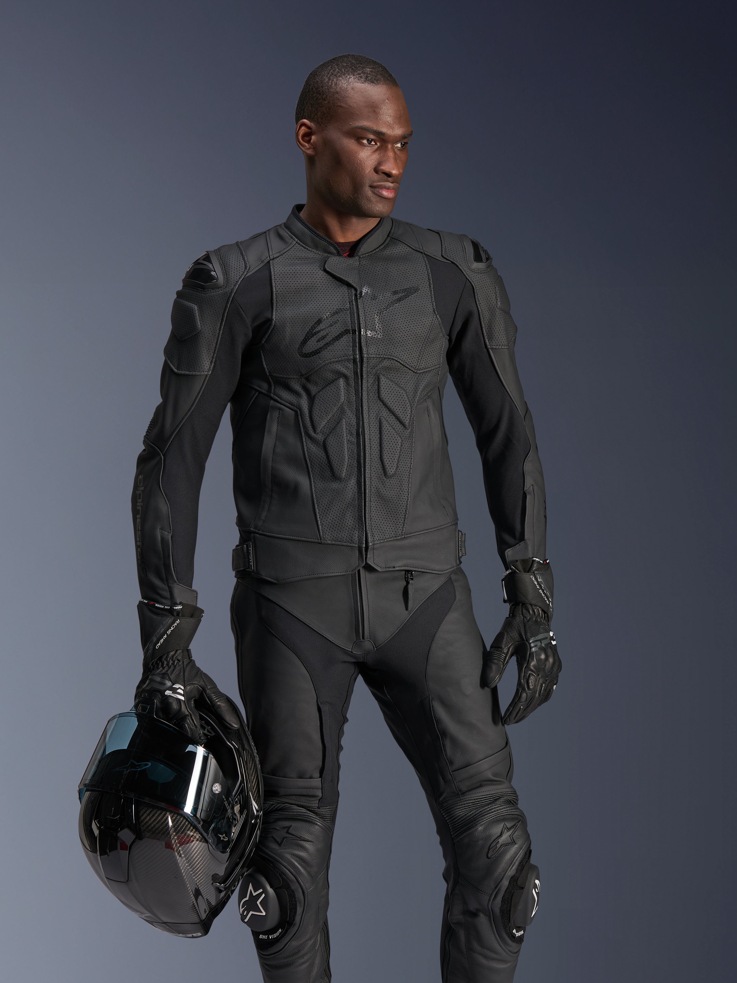 Alpinestars Dusk Airflow läderjacka, svart, bärs av en manlig förare med matchande läderbyxor och handskar, som håller i en svart hjälm, med perforerade läderpaneler och integrerade skydd för sportig motorcykelkörning
