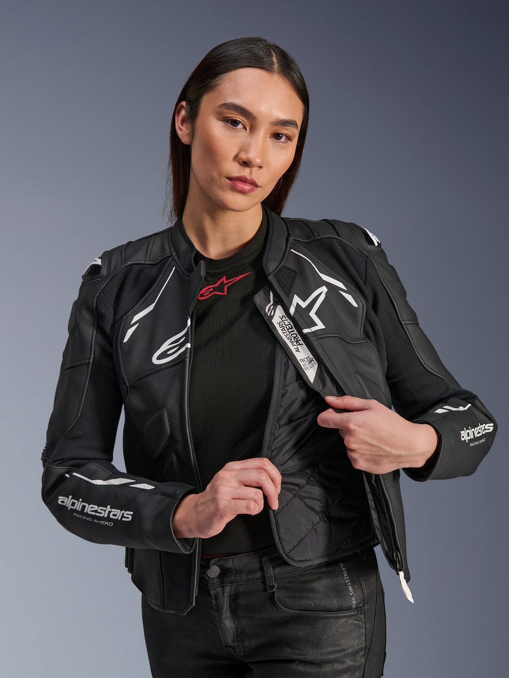 Kvinnlig modell i Alpinestars Stella Dusk läderjacka i svart och vitt, visar det invändiga quiltade termofodret och Tech-Air Ready-etiketten, premium nötläder med vit stjärnlogotyp och 'RACING AHEAD'-grafik på ärmen