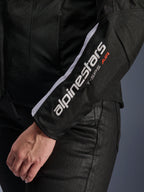Alpinestars Stella T-SPS Air V2-jacka, motorcykeljacka för dam, svart och vit, närbild på ärmen med vit Alpinestars-logotyp och T-SPS AIR-text, mesh- och textilkonstruktion för luftflöde, bärs med svarta mc-byxor