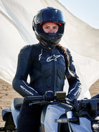 Alpinestars Stella Dusk läderjacka för damer, svart och vit, buren av en kvinnlig förare på en motorcykel med en svart hjälm, med vit logotyp på bröstet och ärmarna, racingfokuserad läderdesign i en utomhusmiljö.