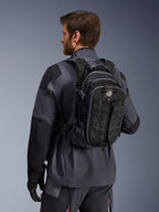 Alpinestars AT-15 ryggsäck, äventyrsryggsäck för motorcykel, svart och mörkgrå, bärs av en förare i en grå jacka, teknisk design med MOLLE-system och 'TERRA'-logotyp, ergonomisk terrängutrustning