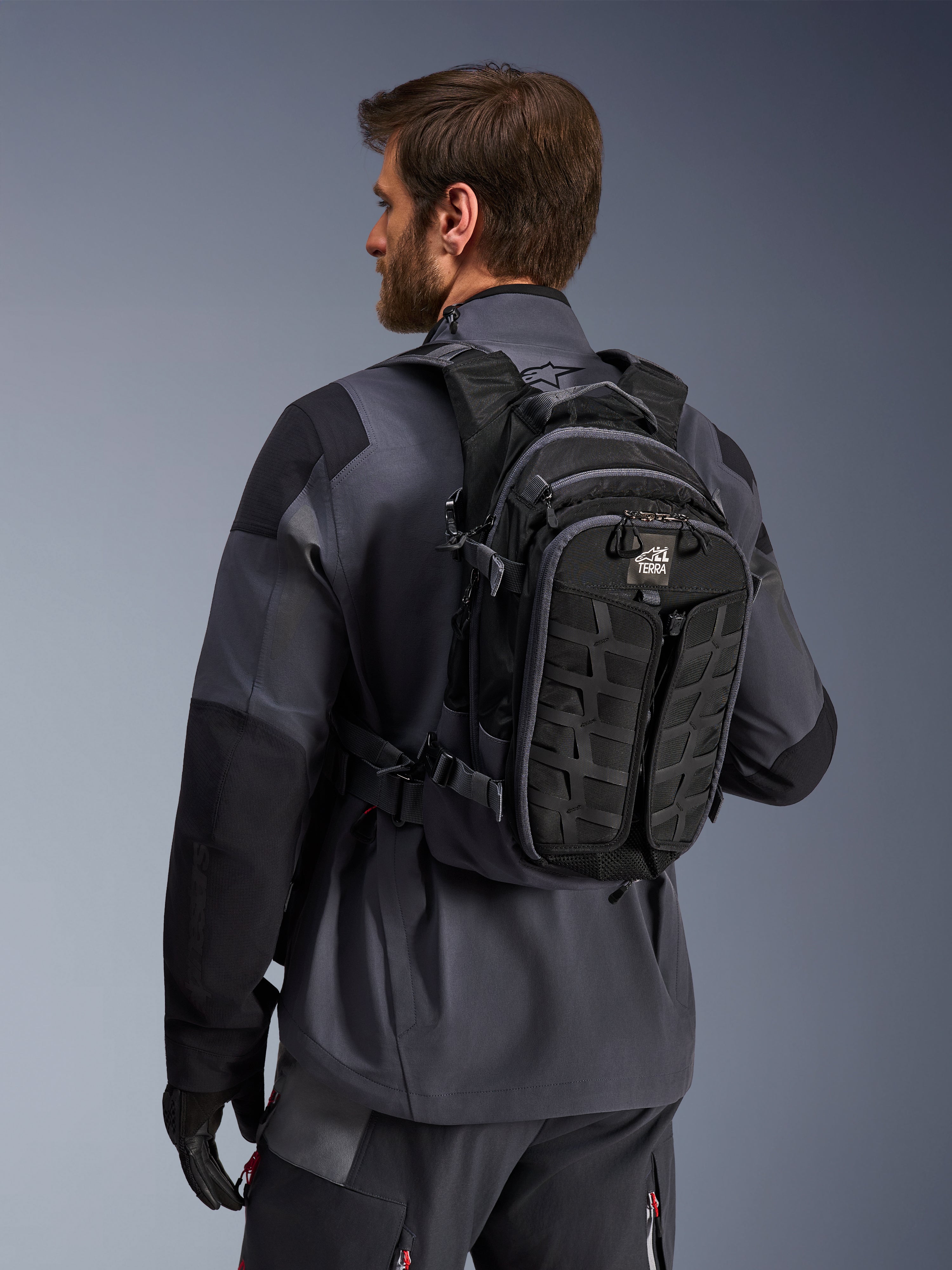 Alpinestars AT-15 ryggsäck, äventyrsryggsäck för motorcykel, svart och mörkgrå, bärs av en förare i en grå jacka, teknisk design med MOLLE-system och 'TERRA'-logotyp, ergonomisk terrängutrustning