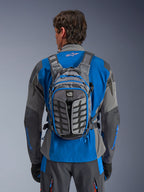 Alpinestars AT-15 Back Pack, äventyrsryggsäck för motorcykel, mörkgrå och djupgrå, bärs av en förare över en blå och grå jacka, med All Terra-branding, ergonomisk strukturell design, justerbara remmar och blå dragkedjsdetaljer.