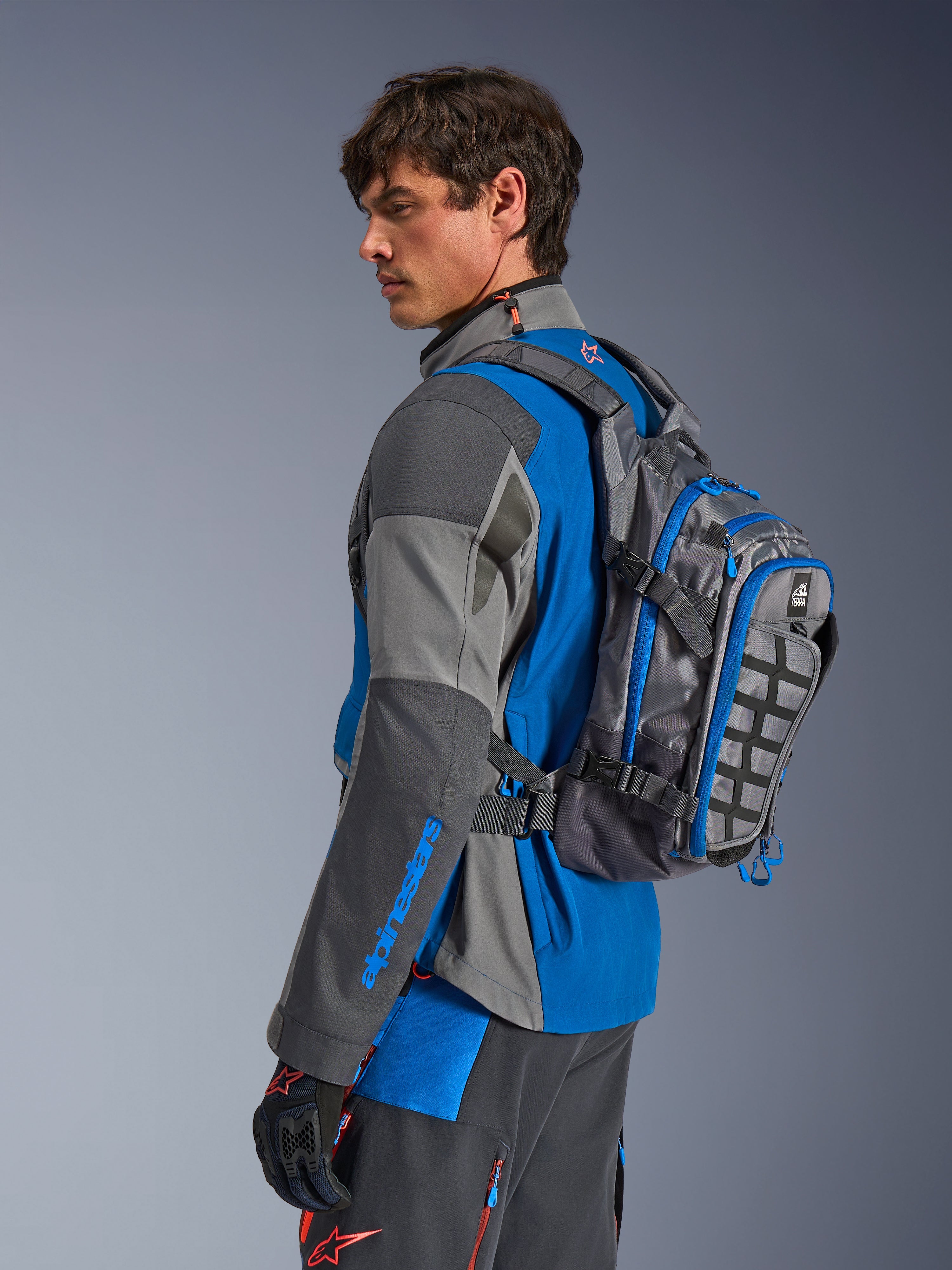 Alpinestars AT-15 Back Pack, äventyrsryggsäck för motorcykel, mörkgrå med blå detaljer, bärs av en förare i matchande grå och blå teknisk körutrustning, utrustad med MOLLE-panel och robust konstruktion för off-road-äventyr.