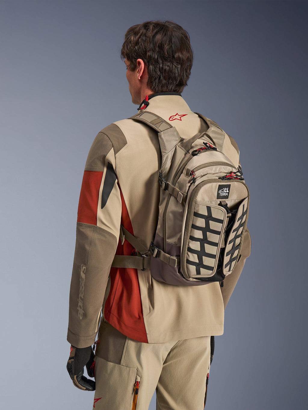 Alpinestars AT-15 ryggsäck i Aluminum Walnut, teknisk motorcykelryggsäck som bärs av en förare i matchande äventyrsutrustning, med beige och brun textilkonstruktion, svarta strukturella detaljer och röda dragtappar, designad för terräng- och äventyrskörning.