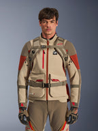 Äventyrsförare iklädd Alpinestars AT-15 ryggsäck i Aluminum Walnut, vy framifrån som visar bröst- och midjeremmar med magnetlås, kombinerat med sandfärgad och röd motorcykeljacka och byxor för äventyr, teknisk terrängutrustning