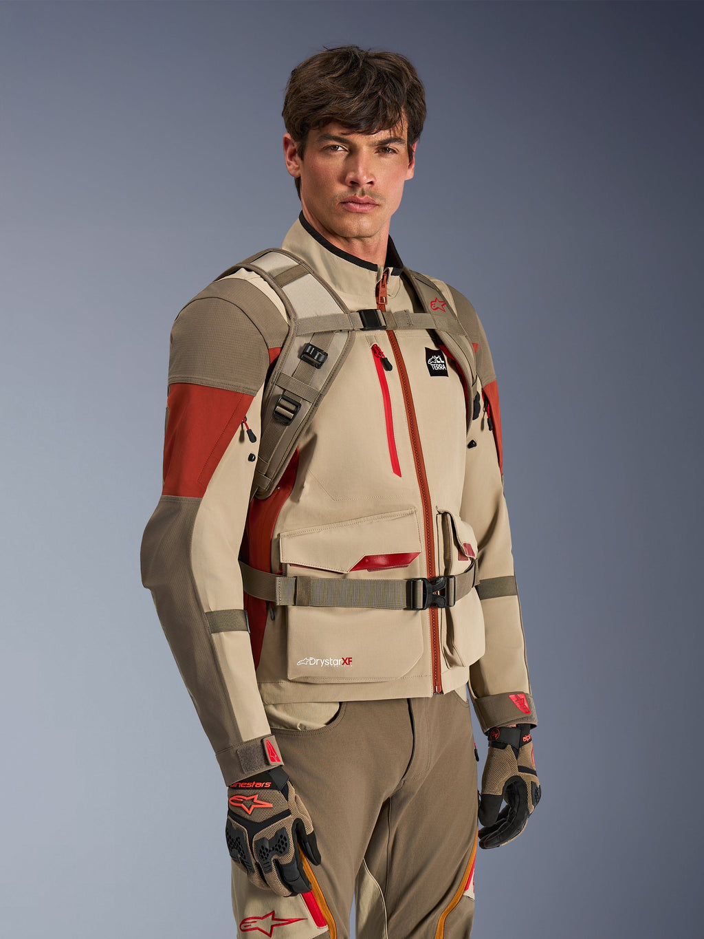 Alpinestars AT-15 ryggsäck i Aluminum Walnut, bärs av en förare över en matchande äventyrsmotorcykeljacka, visar den främre selen med magnetiskt bröstlås och justerbara remmar, beige och röd teknisk utrustning för äventyrskörning