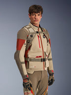 Alpinestars AT-15 ryggsäck i Aluminum Walnut, bärs av en förare över en matchande äventyrsmotorcykeljacka, visar den främre selen med magnetiskt bröstlås och justerbara remmar, beige och röd teknisk utrustning för äventyrskörning