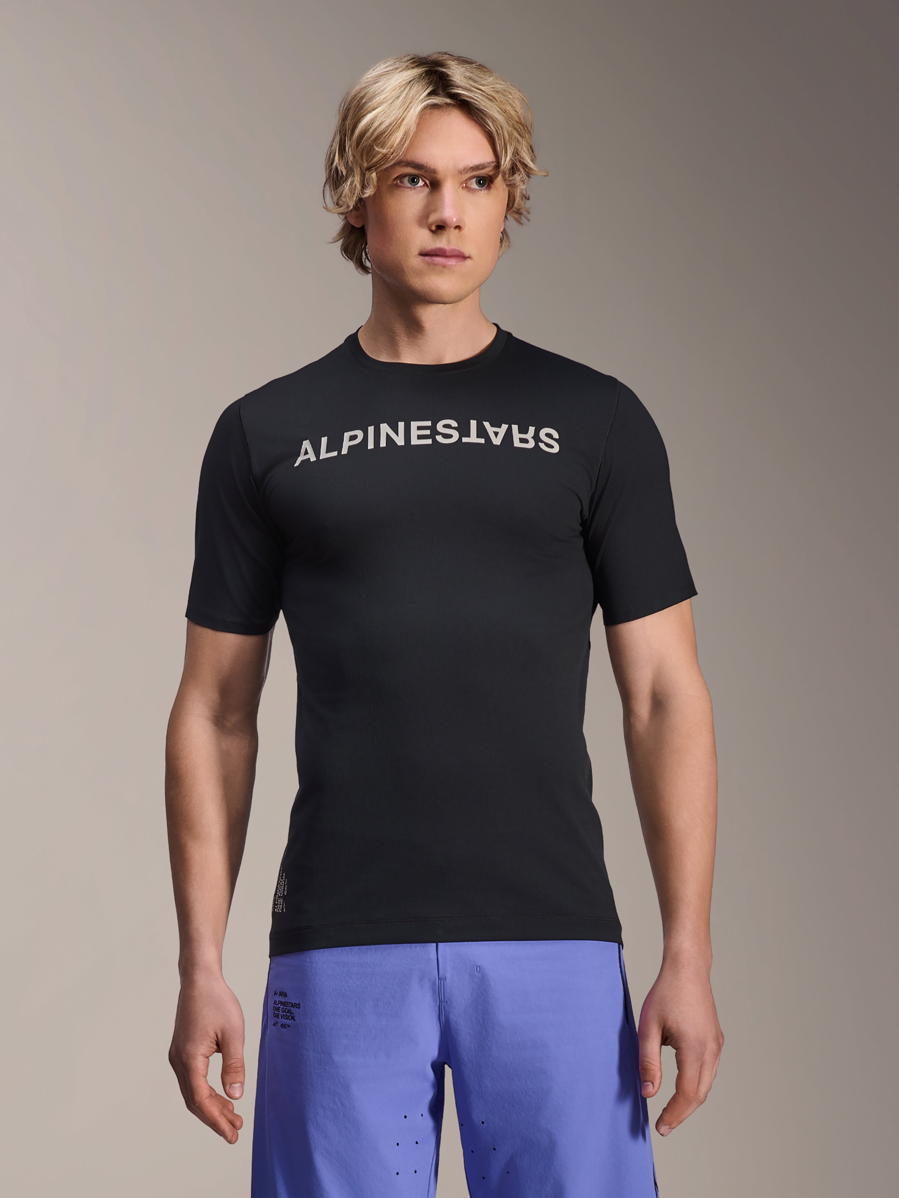 Alpinestars A-Aria Switch kortärmad tröja, MTB-tröja, svart med vit Alpinestars-textlogotyp på bröstet, bärs av en manlig modell med lila shorts, lätt prestandamaterial för terrängcykling