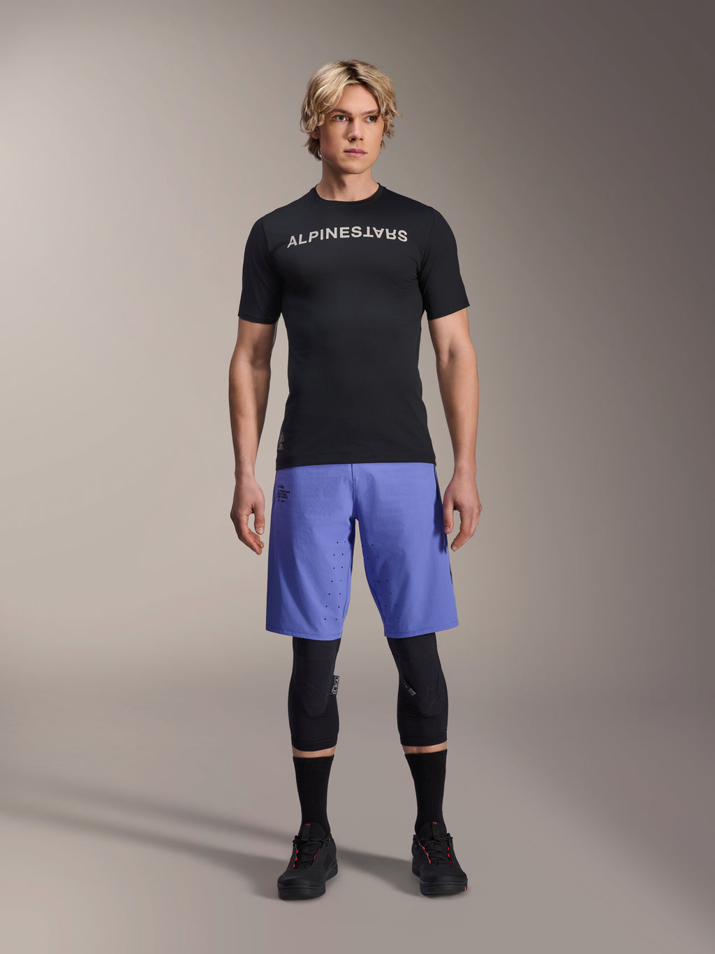 Alpinestars A-Aria Switch-tröja, kortärmad MTB-tröja, svart med spegelvänd vit Alpinestars-logotyp, bärs av en manlig modell med blå cykelshorts och svarta knäskydd, ventilerande funktionskläder för stigcykling
